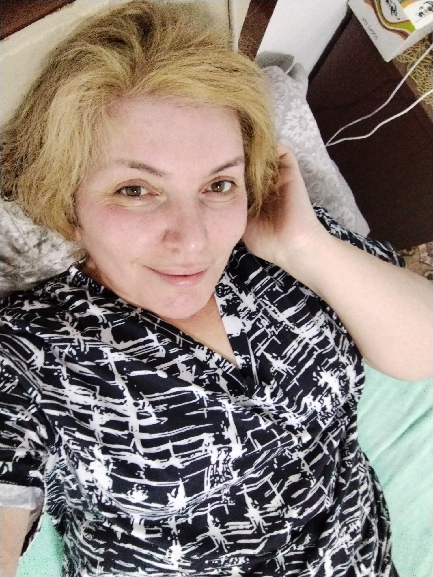 A woman is lying down, facing the camera. She has short blonde hair, light skin, and brown eyes. She is wearing a black and white patterned top. Her right hand is near her head, possibly touching her hair. She is lying on a light green surface, likely a bed. In the background, to the upper right, is a patterned pillow and a box on a wooden surface. The location is not discernable from the image.