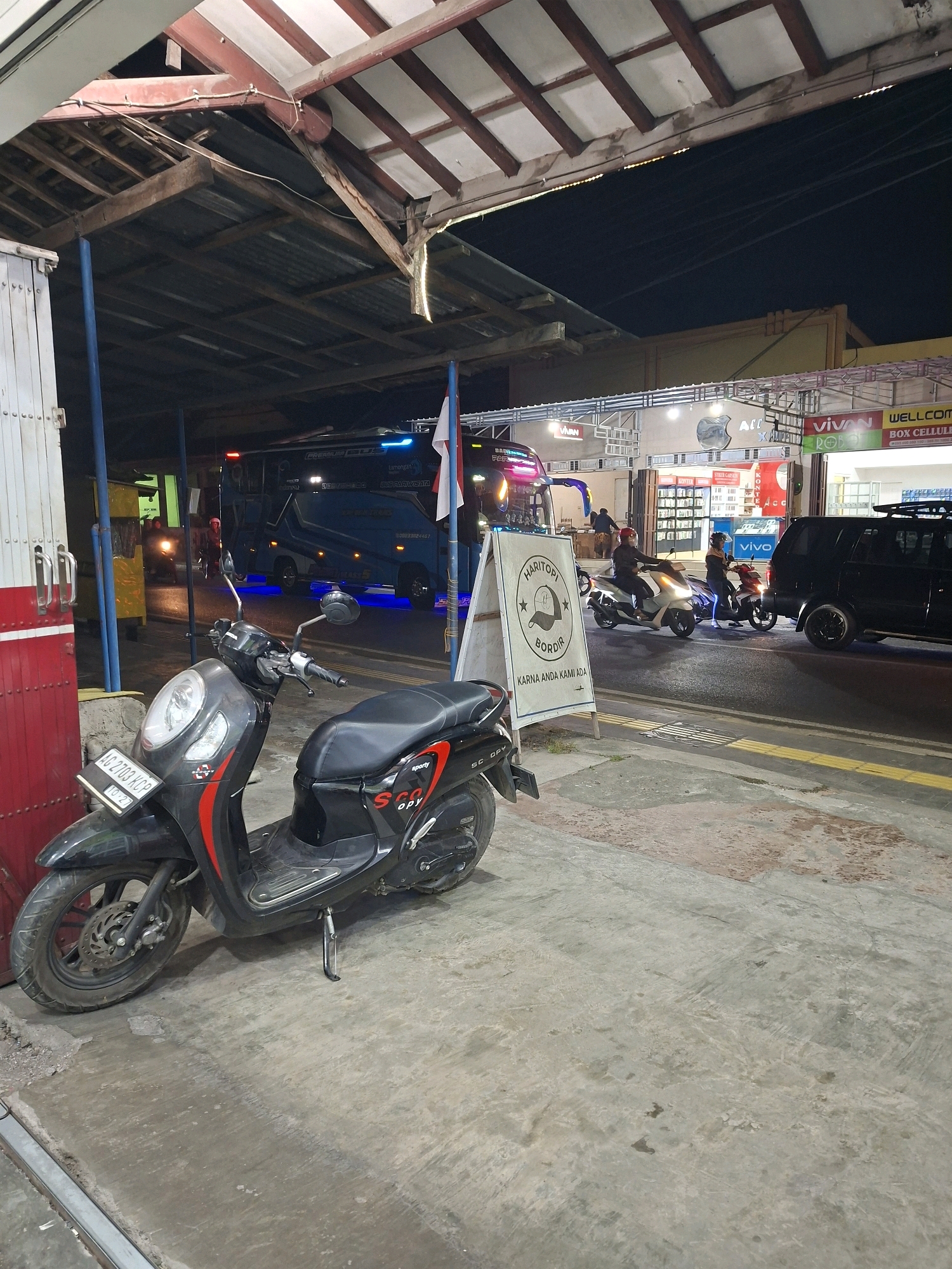 This image depicts a street scene at night in Blitar, Indonesia. In the foreground, a black Honda Scoopy motorcycle is parked on a concrete surface. The motorcycle features red and black accents and has a license plate that reads "AG 2703 KCP". Behind the motorcycle, a blue bus with illuminated blue lights along its side is stopped, likely at a bus terminal or stop. The bus displays the text "PREMIUM BUS". Further back, a black SUV is parked on the right side of the road.
Several people are visible: two individuals are riding on a scooter in the middle ground, one wearing a red helmet and the other a white helmet. Another person is partially visible on a different scooter. In the background, there are commercial buildings with signs, including "WELLCOME", "VIVAN", and "BOX CELLULER", suggesting a commercial area. A white A-frame sign with text that appears to be in Indonesian is placed in front of the bus. The scene is illuminated by artificial lights from the buildings and the bus. The overall environment suggests a somewhat urban setting at night.