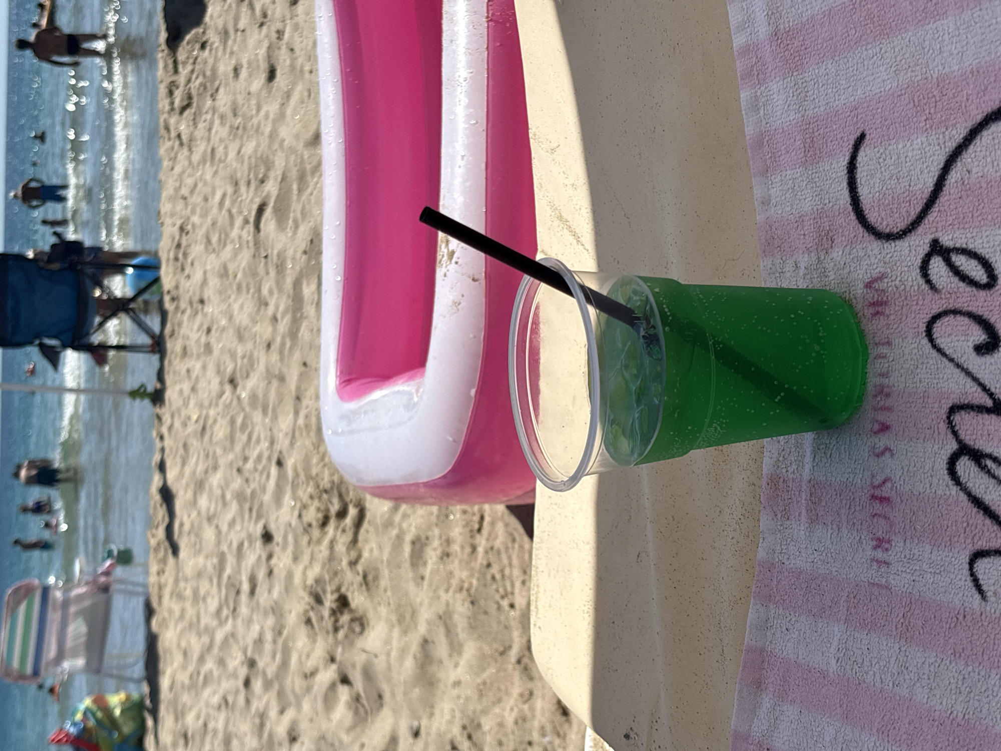 The image shows a bright, sunny day at a beach in Primorsko, Bulgaria. In the foreground, a green beverage with a straw sits in a clear plastic cup on top of a towel with pink and white stripes and black script lettering. Behind the drink, a pink and white inflatable pool is filled with water. The beach sand stretches out behind the pool, and further in the distance, people are visible in the blue ocean, some wading and others swimming. Beach chairs and umbrellas can be seen on the sand, suggesting a recreational area. The overall scene conveys a relaxed, summery atmosphere.