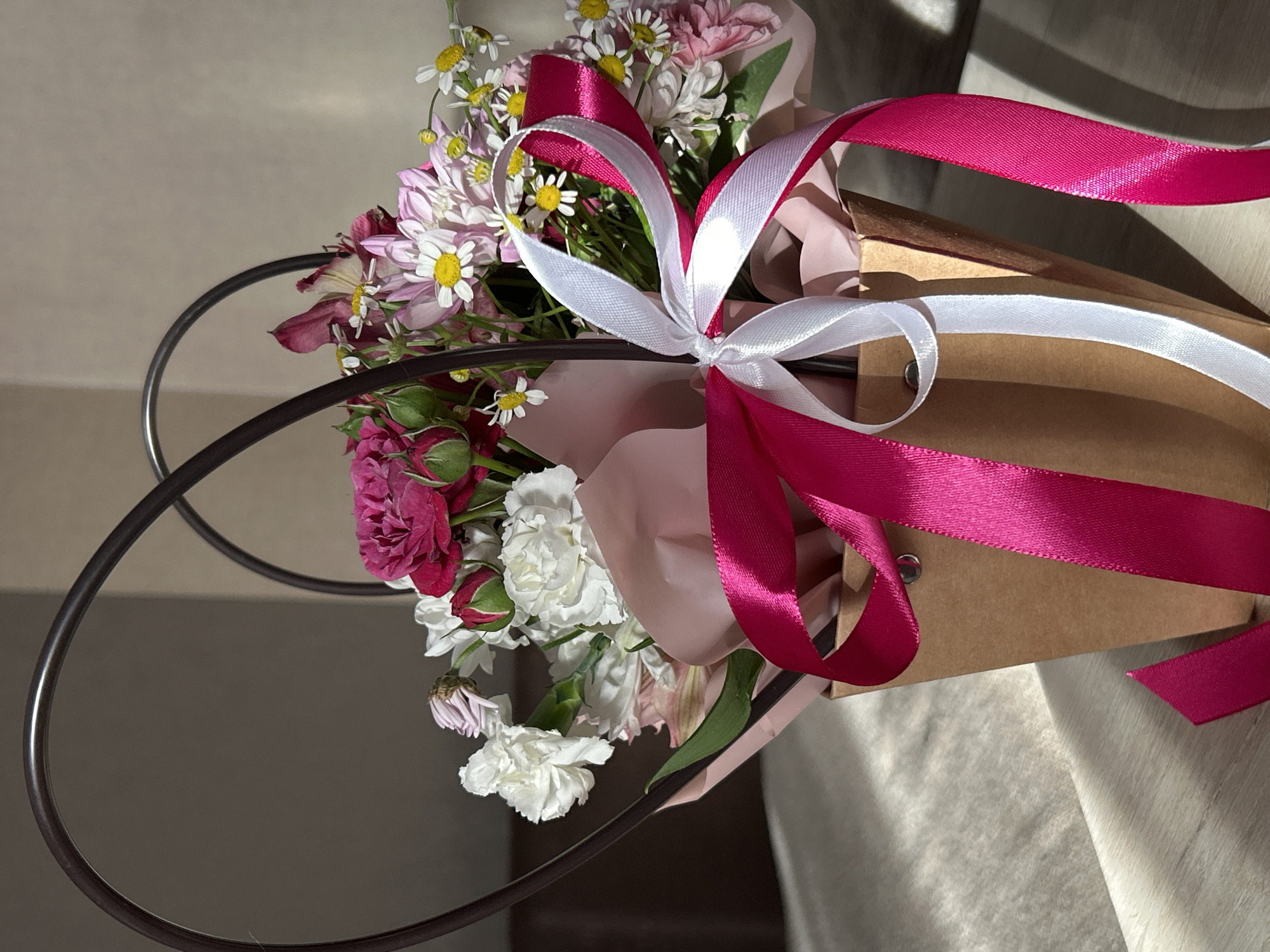 A vibrant bouquet of mixed flowers is prominently featured, likely presented as a gift or for a special occasion. The arrangement includes pink and white daisy-like chrysanthemums with yellow centers, small fuchsia roses and rosebuds, and ruffled white flowers resembling carnations. These flowers are artfully wrapped in light pink paper and housed within a simple brown craft paper bag or box.
A decorative element is a striking satin ribbon, with both white and fuchsia strands, tied in an elaborate bow around the bouquet, enhancing its presentation. In the foreground and midground, a dark, curved, metallic-looking object, possibly a handle for the container or a decorative frame, adds a structural element to the composition.
The scene is illuminated by natural light, casting soft shadows and highlighting the textures of the flowers and ribbons, suggesting it was captured during daytime. The bouquet rests on a light, textured surface, possibly fabric, against a plain, light-colored background. There is no visible text in the image. The overall impression is one of freshness and thoughtful arrangement. The location context is Марковский, Russia.