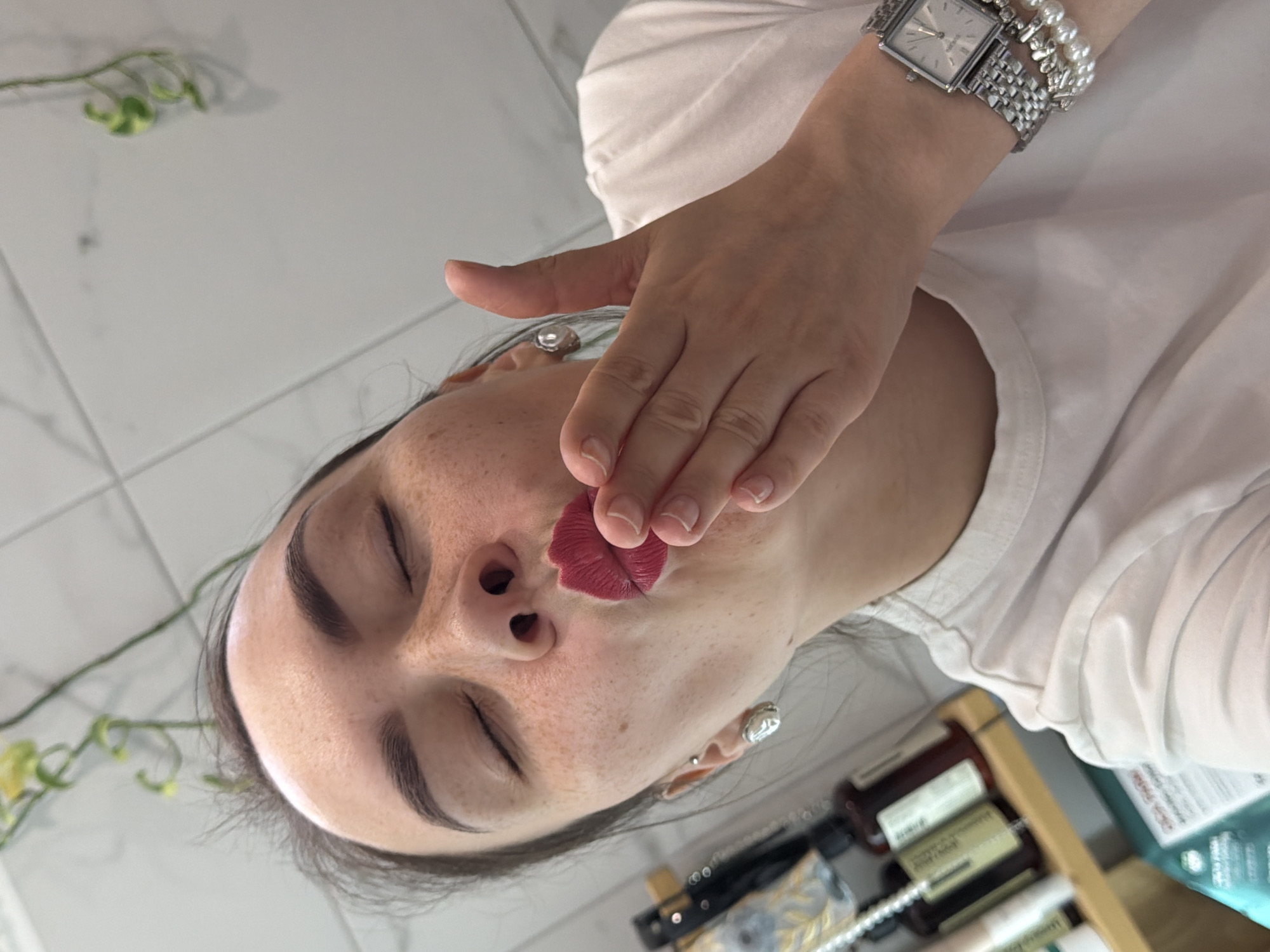 A woman with freckled skin and closed eyes, wearing a white t-shirt, is holding a lipstick transfer in the shape of lips to her face. She has a silver watch and bracelet on her wrist, and silver earrings in her ears. The background includes a white tiled wall, a green plant, and various bottles on a shelf. The location is Chaikovsky, Russia.