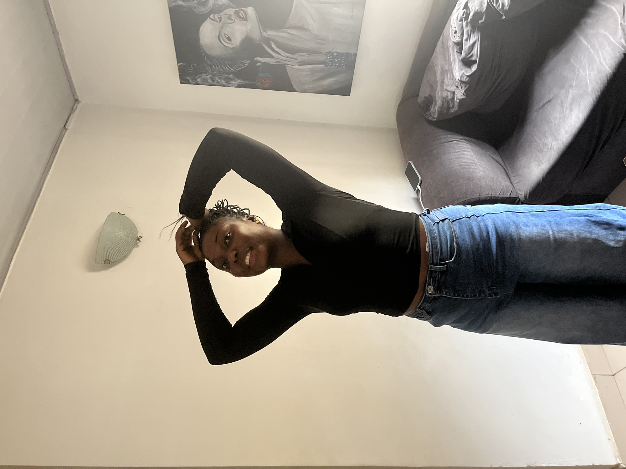 A young Black woman with her hair pulled up in a bun stands in a room, touching her hair with both hands. She is wearing a black long-sleeved shirt and loose-fitting blue jeans. The room has white walls, a light fixture on the ceiling, and a large painting of a woman on the right wall. A dark gray sofa is visible in the background, with a phone charging on its surface. The scene appears to be an indoor residential setting in Abuja, Nigeria.