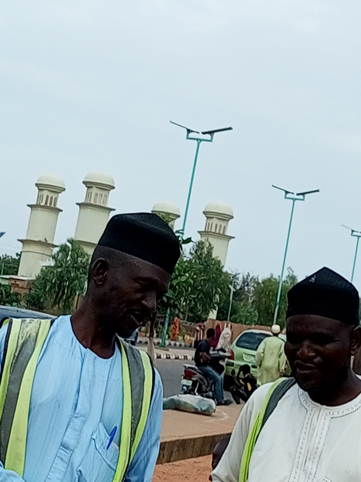 The image depicts a street scene in Gombe, Nigeria, with two men in the foreground wearing traditional Islamic caps and vests. The man on the left wears a light blue striped garment and a lime green and yellow safety vest. He has a pen in his chest pocket. The man on the right wears a white embroidered garment and a lime green vest. In the background, several minaret-like structures, possibly part of a mosque or religious complex, are visible against a cloudy sky. Streetlights with solar panels are also present. Other people and vehicles, including a motorcycle and a car, are visible further down the street, suggesting a moderately busy urban environment. The ground appears to be paved with some dirt and concrete elements.