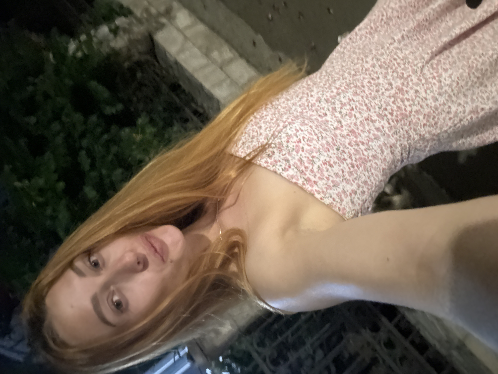 A woman with long, red hair and fair skin is posing for a selfie outdoors at night. She is wearing a pink floral print dress with a sweetheart neckline. The background shows greenery and some stone structures, possibly part of the city of Budva, Montenegro, with artificial lighting illuminating the scene.