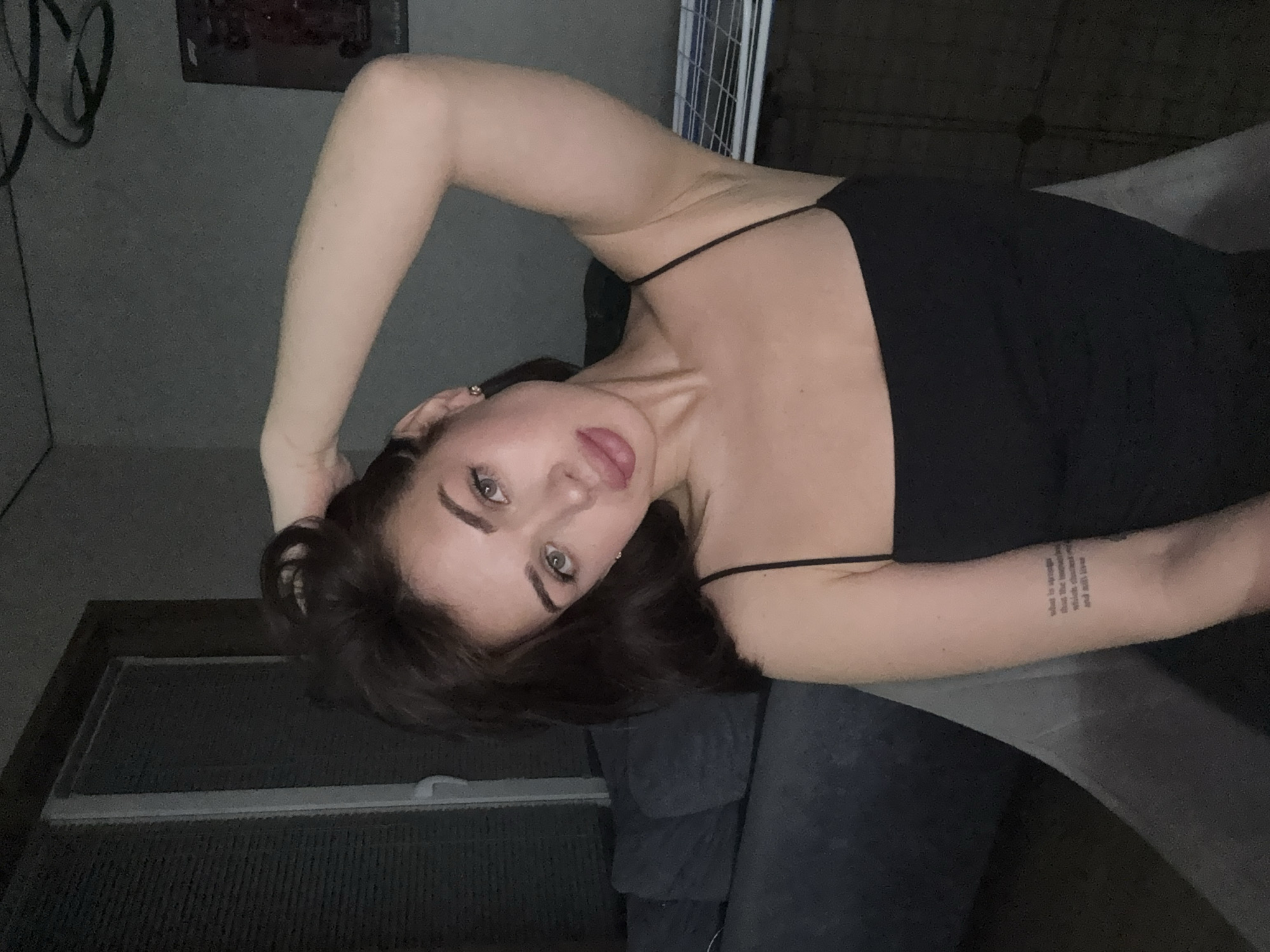 A woman with dark hair and fair skin is reclining on a gray upholstered surface, possibly a sofa. She is wearing a black top with thin straps. Her left arm is bent and raised above her head, while her right arm rests on a clear surface in front of her. She has a tattoo on her right forearm. The background includes a dark-colored wall, a window with blinds, and a wire rack. An indistinguishable framed image is on the wall. The location is specified as Лобаново, Russia.