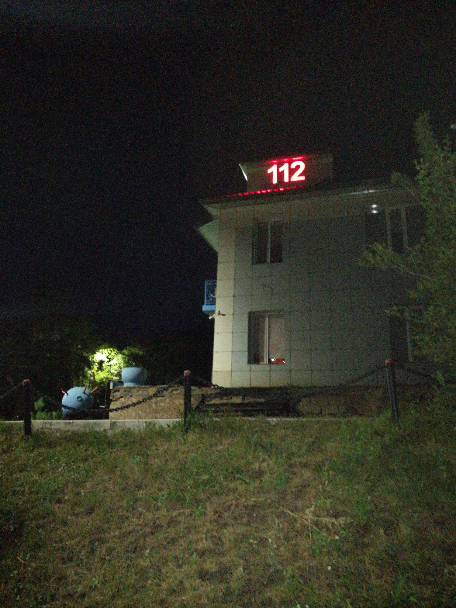 This is a nighttime, low-light, grainy photograph of a building in Pavlodar, Kazakhstan. The building is a two-story structure with a light-colored facade, possibly concrete or stucco. A prominent red neon sign displaying the number "112" is mounted on the roof of the building. The building has multiple windows, some of which are illuminated from within, and a blue balcony or railing is visible on the upper level.
In the foreground, there is a grassy area with scattered vegetation. Behind the grass, a low concrete wall or embankment is visible, topped with a chain fence. On the left side of the embankment, a blue, spherical object with protrusions that resemble eyes and antennae is situated. This object appears to be a decorative element or a character statue. A chain is draped over the embankment and connected to posts supporting the fence. To the right of the blue object, a second, lighter-colored spherical object is partially visible. Trees and foliage are present in the background, illuminated by artificial light sources. The overall environment suggests a semi-urban or roadside location.