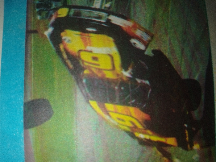 A yellow, black, and red race car, marked with the number 19, is depicted in a dynamic, sharply tilted position on a race track in the city of Unknown, Unknown. The vehicle appears to be on two wheels or in a state of partial air during a high-speed maneuver or incident. The car's underside is partially exposed due to its lean. The surrounding environment includes a green grassy area adjacent to the asphalt track. An indistinct dark object, possibly a tire or part of another vehicle, is visible in the upper left background. No individuals are present in the frame. The image quality is low, characterized by blur and grain.