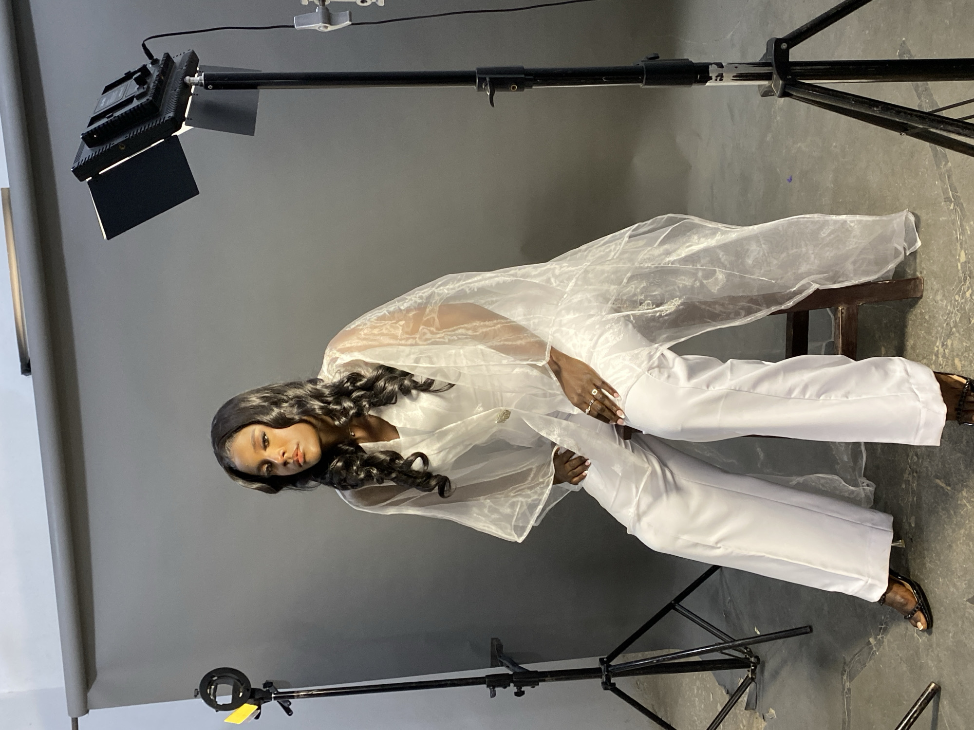 The image depicts a woman posing in a studio. She is wearing a white pantsuit with a sheer white overlay. She is seated on a wooden stool, with a gray backdrop and studio lighting equipment visible. There are no indications that this image was taken in Jos, Nigeria or any other particular city; this is a studio shot. The scene includes lighting stands, a ring light, and other studio equipment.