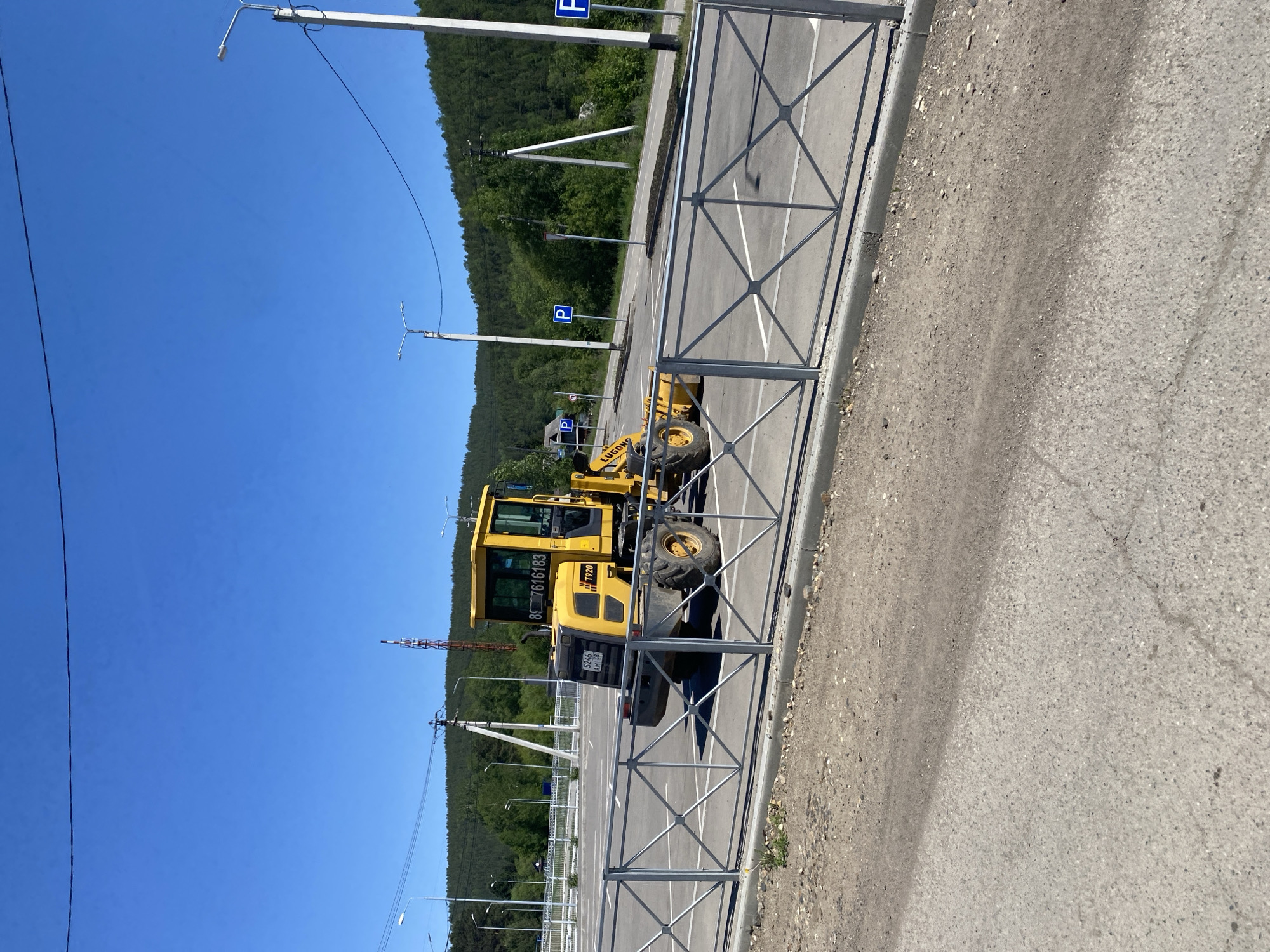 A yellow wheel loader with the brand "LUGONG" and model "T920" is parked in an empty parking lot under a clear blue sky. The vehicle has visible license plates. The parking lot is delineated by white lines and surrounded by a metal fence. In the background, a dense forest covers rolling hills. Several streetlights and utility poles are also present. The scene is set in the city of Bolshoy Lug, Russia.