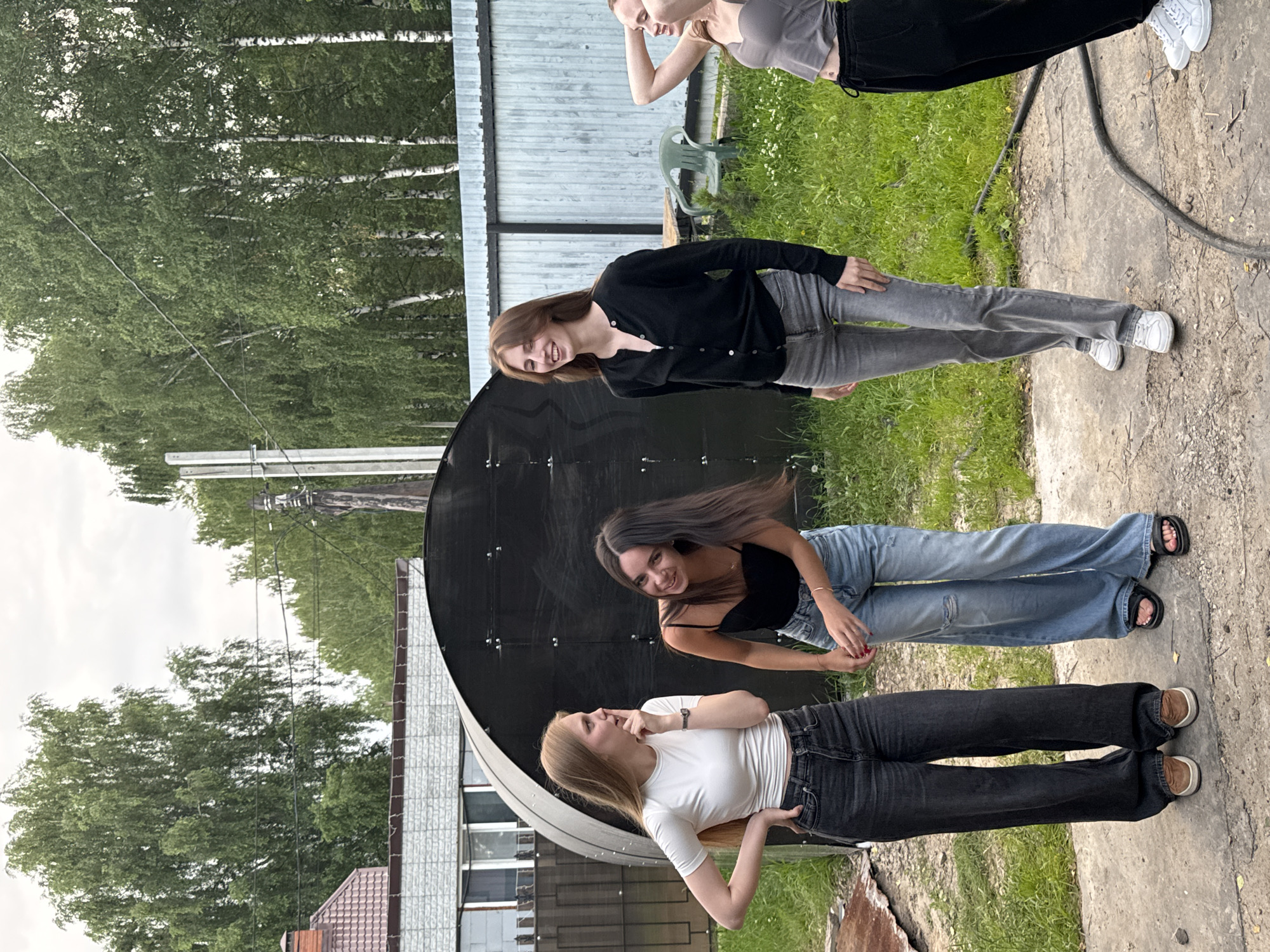 In an outdoor setting with lush green grass and trees, four young women are gathered. The scene appears to be a backyard or a similar casual outdoor space, possibly during daylight hours with overcast skies. The women are dressed in casual attire, with two of them wearing jeans and t-shirts or sweaters, and the other two in jeans and tank tops.
One woman, with long brown hair, is smiling and looking towards the camera, wearing a black tank top and light blue distressed jeans. Another woman, with blonde hair, is also looking at the camera and touching her nose, dressed in a white t-shirt and black jeans. A third woman, also with long brown hair, is wearing a black cardigan over a white top and grey jeans. The fourth woman, at the top of the frame, is partially visible, wearing a grey tank top and black pants.
The women are positioned around a large, black, semi-circular object, possibly a piece of equipment or a structure, with a corrugated metal fence in the background. The ground is a mixture of concrete and dirt, with some loose gravel. The overall mood seems relaxed and informal, suggesting a casual get-together or photoshoot. There is no visible text in the image. The location context provided is Perm, Russia.