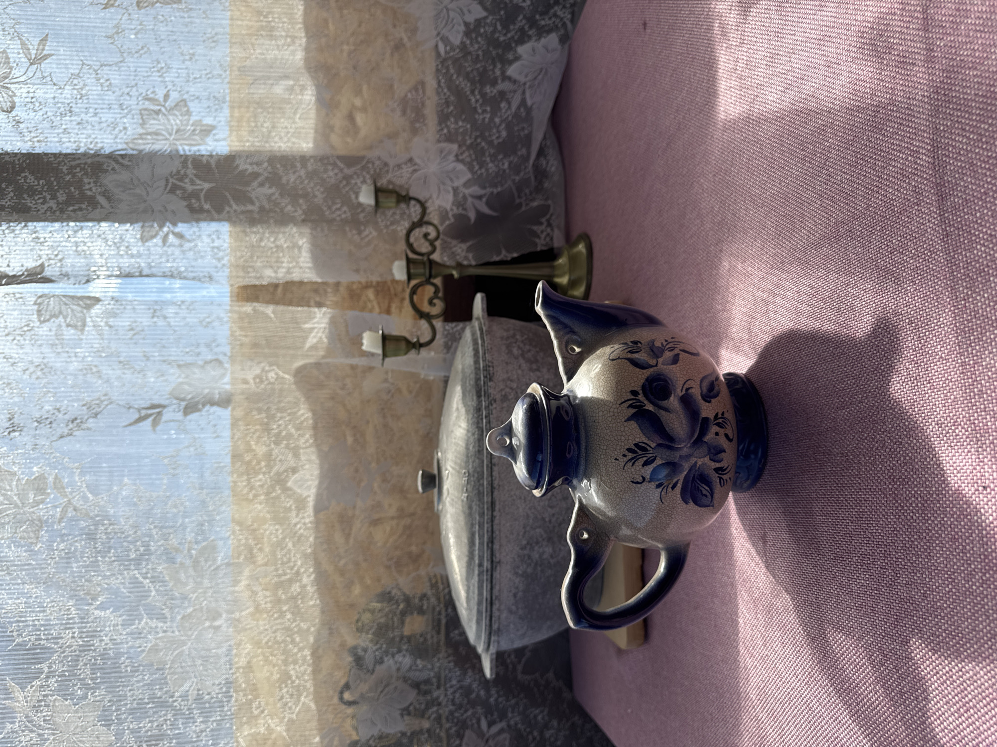 The image depicts a still life scene indoors. A floral patterned curtain is on the left, and a lilac-colored textile surface is on the right. An aged pewter cooking pot with a lid and a ceramic pitcher with blue floral designs are positioned on the lilac surface, in front of the curtain. A golden candelabra stands behind the cooking pot. The scene is lit by natural light, casting shadows across the items. There is no information to confirm that the location is in the city of Chaikovsky, Russia.