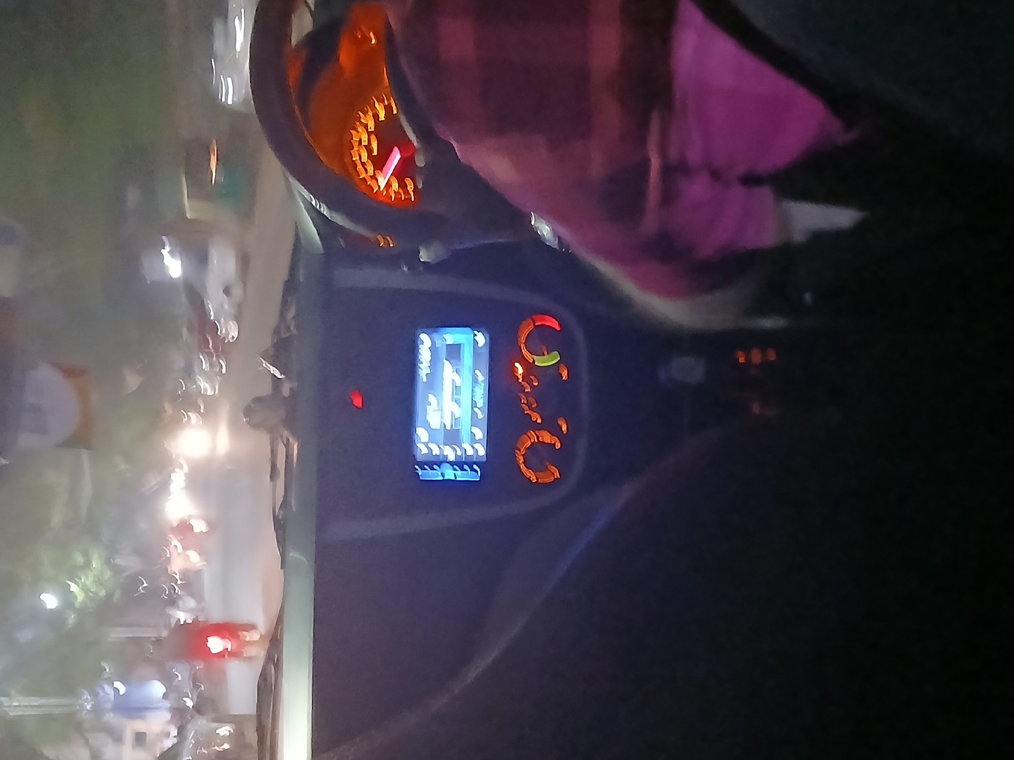 The image depicts an interior view from within a vehicle at night, likely a car. A blurred, orange-lit speedometer and digital display are visible on the dashboard. Part of a person's upper body, wearing a pink and patterned shirt, is present. Outside the vehicle, through the windshield, there are blurry lights suggestive of a street scene, including a possible traffic signal. Given the prompt, it is inferred that the location is Bengaluru, India. The details are obscured due to the low-light conditions and motion blur.