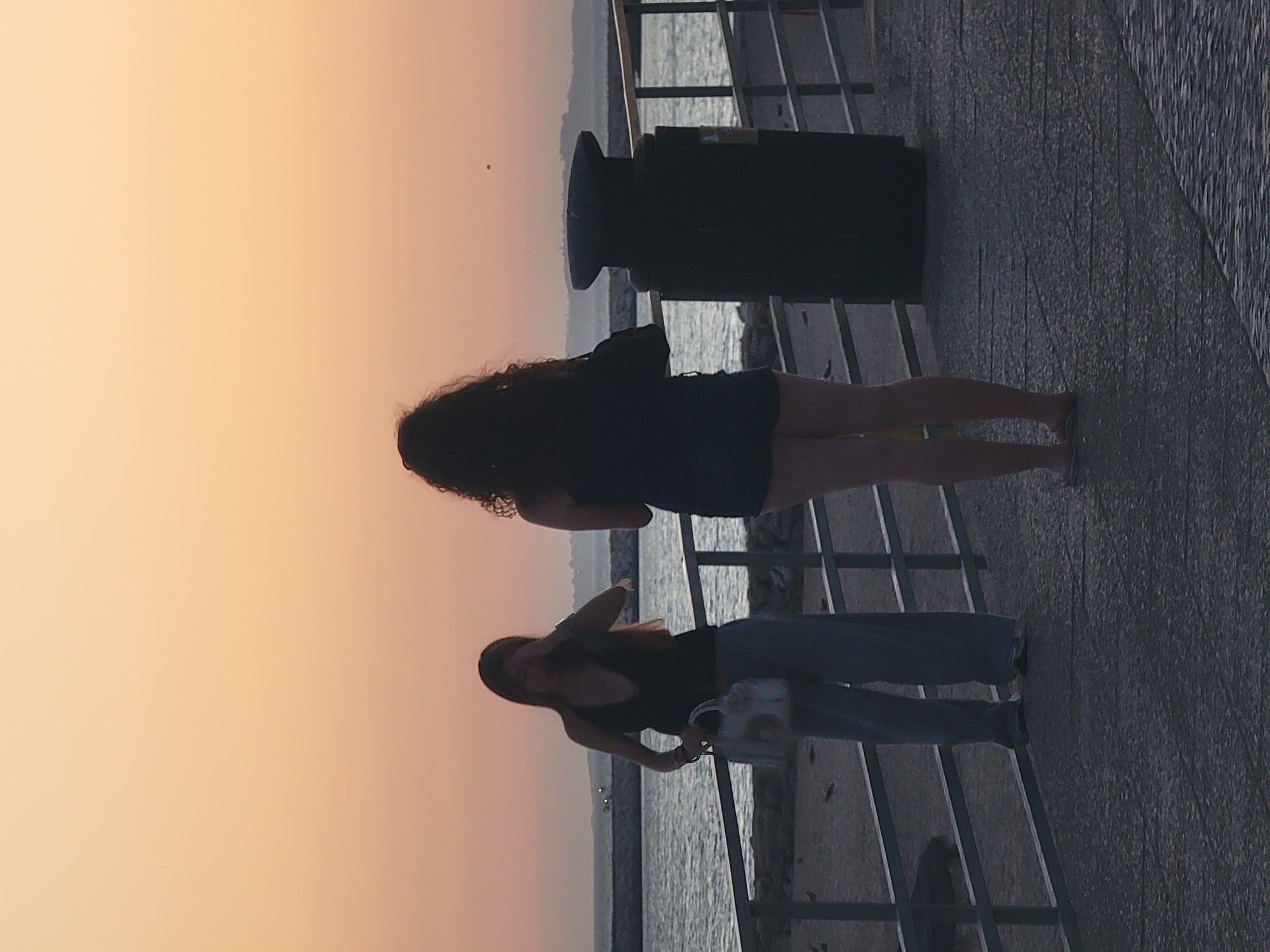 Two women stand on a promenade in Quarteira, Portugal, near the Atlantic Ocean, during a sunset. The sky exhibits a gradient of orange and pink hues. On the left, a woman with long brown hair wears a dark halter top and wide-leg light blue pants, holding a white handbag. On the right, a woman with curly dark hair, seen from behind, wears a dark sleeveless top and dark shorts. A large dark cylindrical trash bin is positioned between them on the promenade. The promenade is paved with patterned grey stones and bordered by a metal railing. Beyond the railing, the sea is visible with gentle waves and a distant stone pier. The overall lighting suggests late afternoon or early evening.