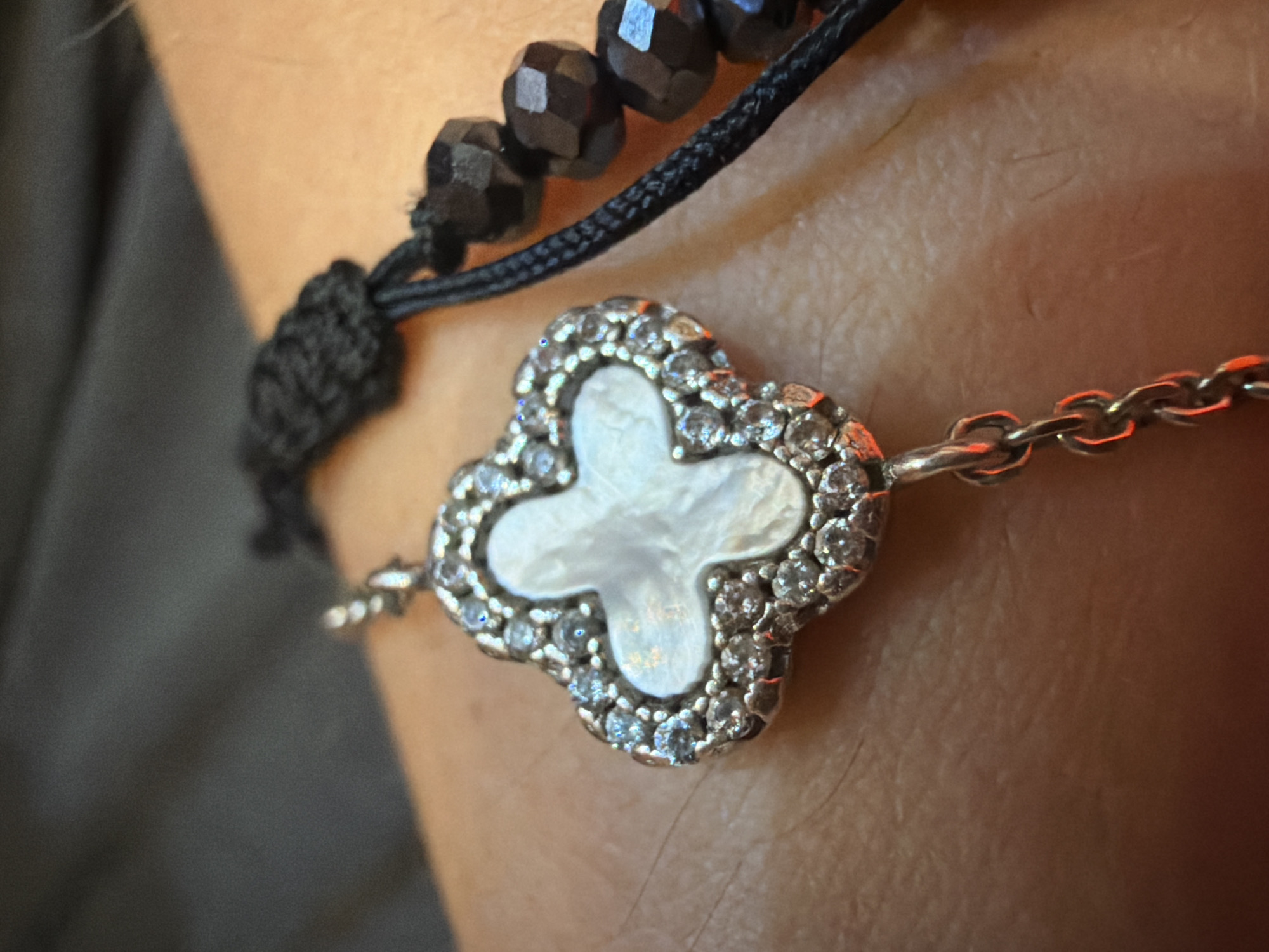A close-up shot shows a four-leaf clover pendant with a mother-of-pearl inlay and a halo of small, sparkling stones, possibly diamonds or cubic zirconia. The pendant is attached to a delicate silver chain. To the right of the pendant, a black string bracelet is visible, adorned with faceted dark beads. The jewelry rests on a person's skin, which appears to be an arm or wrist, with fine hairs visible. The background is softly blurred, showing dark fabric and the skin's texture. The lighting creates reflections and highlights on the jewelry, giving it a luminous quality. The scene is devoid of any specific location markers within a city, making its setting unknown.