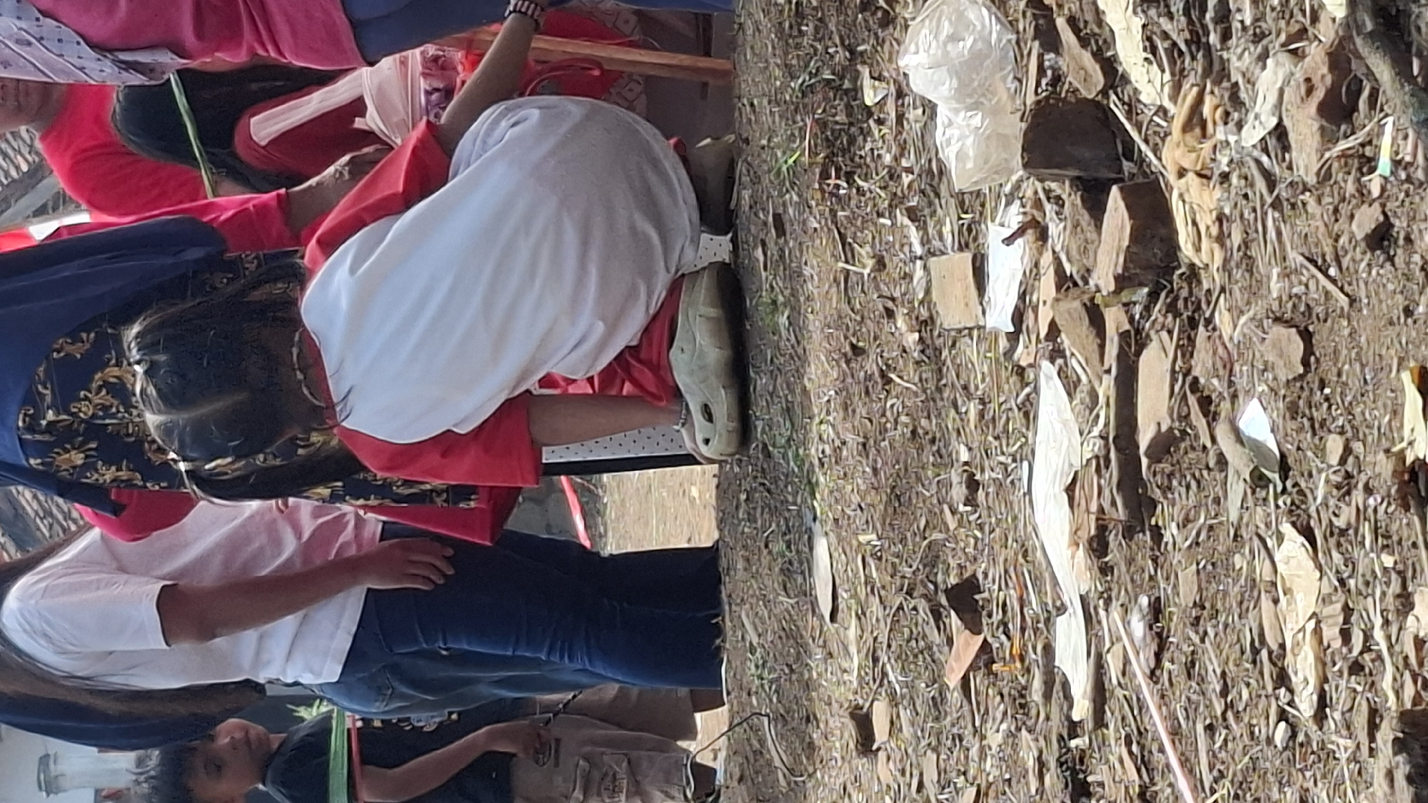 The image depicts a group of people in an outdoor setting. In the foreground, a young child with dark hair styled in pigtails is crouching down, wearing a red and white shirt and light-colored shoes. To the left, a young boy in a black shirt and tan shorts is visible. Behind the children, several adults are standing. One woman is wearing a white t-shirt and blue jeans. Another woman is wearing a blue patterned garment and a headscarf. Further in the background, other individuals in red shirts are present. The ground is covered with dirt, debris, and scattered pieces of what appear to be bricks or rocks. The overall environment suggests a public gathering or an event, possibly in a somewhat unkempt area. The location is identified as Cilaku, Indonesia.