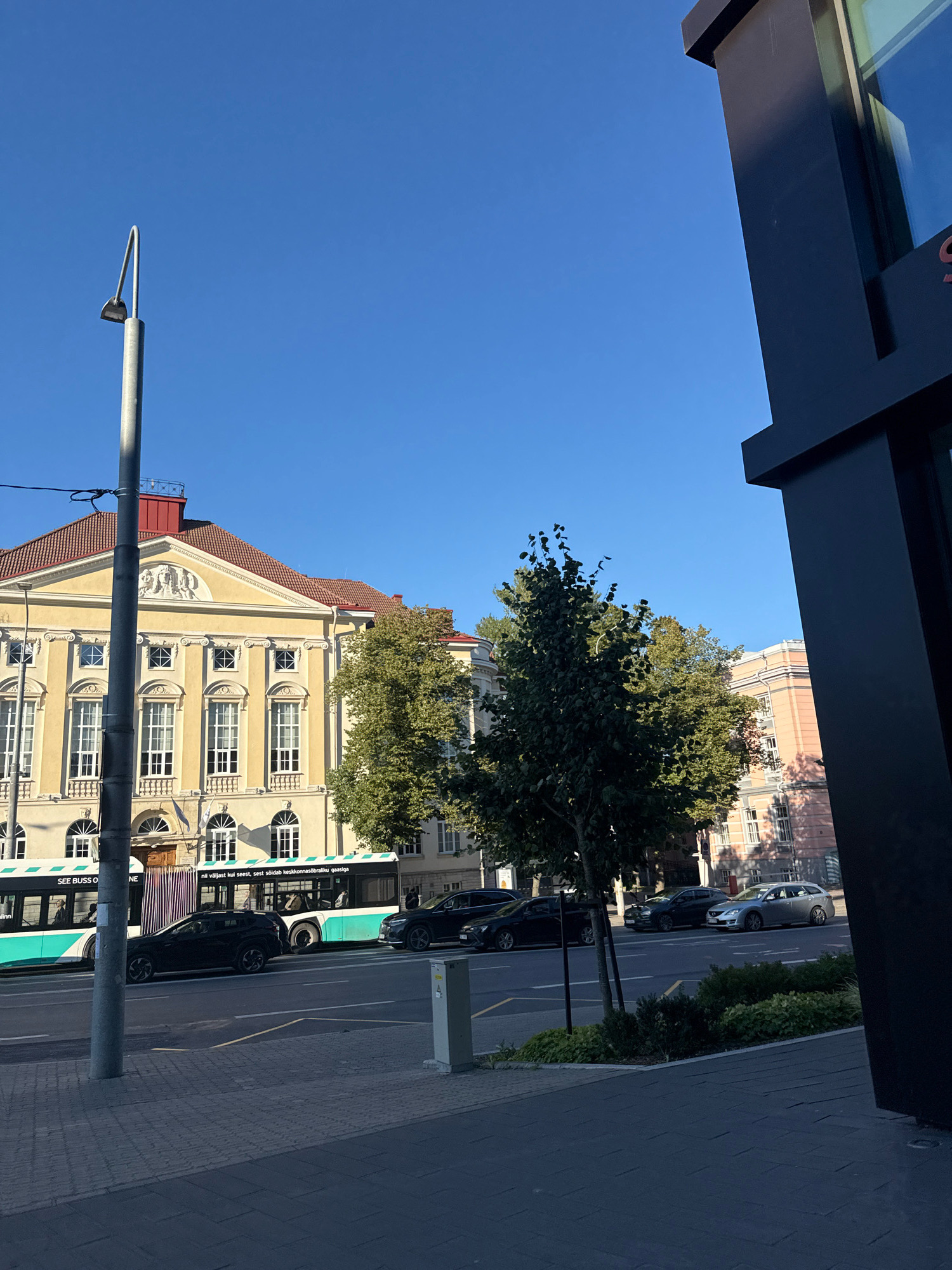 The image captures a street scene in Tallinn, Estonia, on a clear, sunny day. In the foreground, a gray tiled sidewalk is visible, bordered by a strip of green plants and bushes. A light gray utility box stands on the sidewalk. A lamppost with a curved arm is positioned on the left side of the image.
The street features several vehicles. A turquoise and white bus with visible passengers is in the left lane, and a black SUV is in the right lane, moving away from the camera. Further down the street, several other cars are parked or in motion, including a silver station wagon.
In the background, a large, ornate yellow building with a red tiled roof stands prominently. The building has multiple windows, some with decorative white frames, and architectural details such as columns and a sculpted frieze above the entrance. To the right of the yellow building, a lighter pink building with white window frames is partially visible. Trees with green foliage are scattered throughout the scene, with a prominent tree in the mid-ground. A dark, modern building structure dominates the far right of the image, casting a shadow. The sky above is a clear, bright blue.