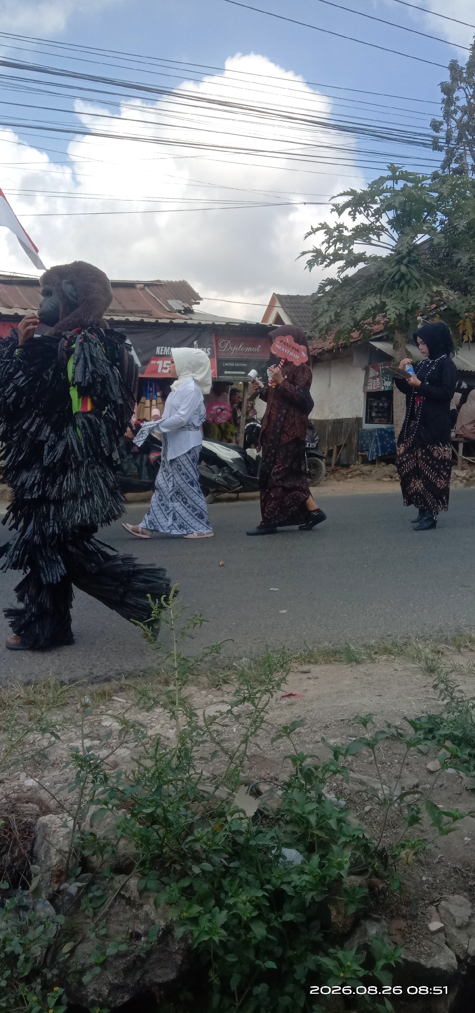 In Pakong, Indonesia, a parade is underway on a paved street. The procession features a person in a gorilla costume, its body covered in black, shaggy material, walking alongside three women. Two of the women are dressed in traditional batik attire. One woman in a white headscarf and a blue and white patterned sarong walks beside the gorilla, while another woman in a patterned brown dress holds a red sign that reads "PANTANG MENYERAH" (Never Give Up). The third woman, also in traditional attire, walks slightly behind and to the right. Buildings line the street, and overhead, power lines crisscross against a cloudy sky. The scene suggests a community event or celebration.