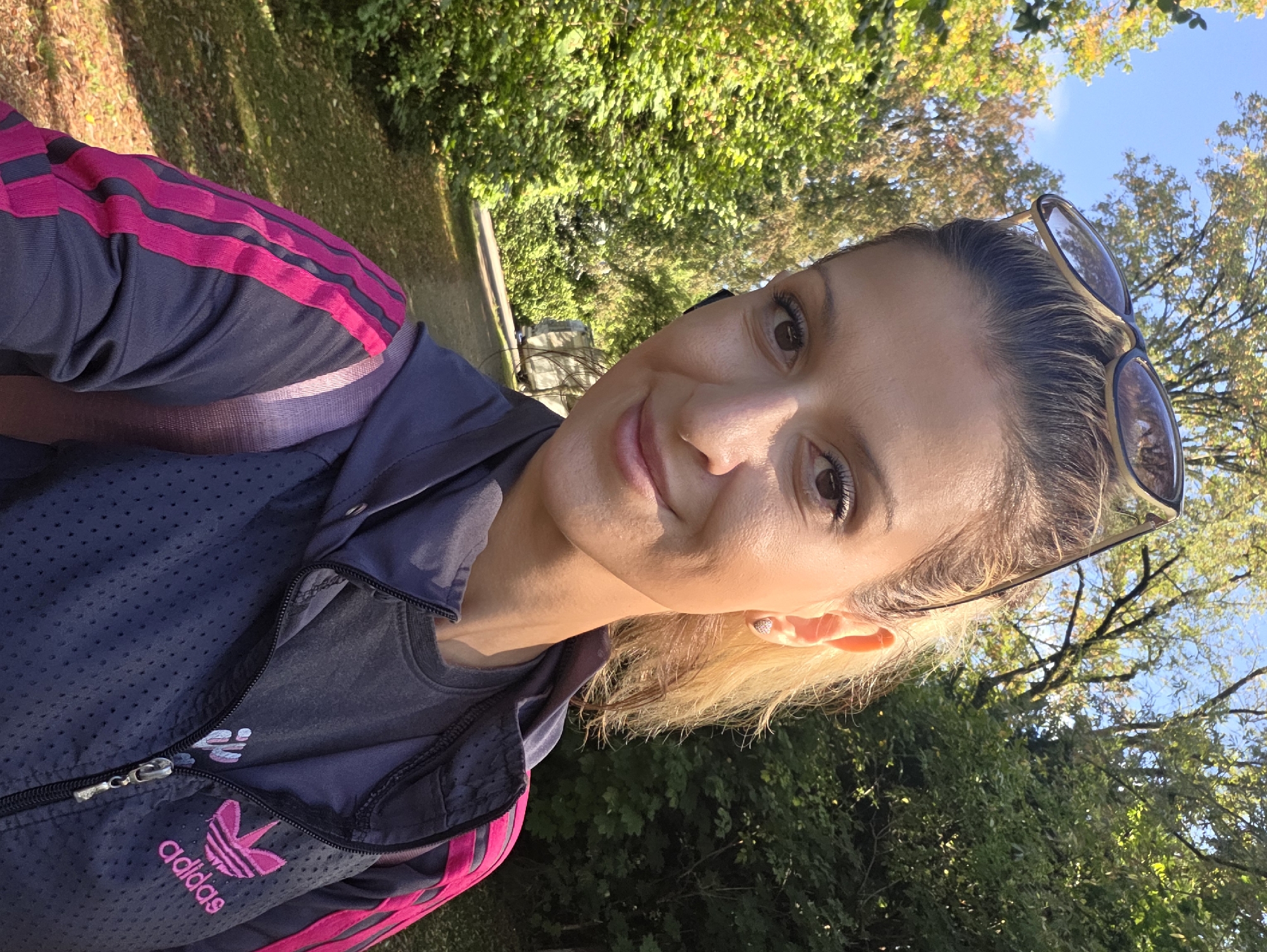 In Munich, Germany, a woman with blonde hair pulled back in a ponytail and sunglasses perched on her head takes a selfie outdoors. She is wearing a dark gray Adidas jacket with hot pink stripes down the sleeves and a matching hot pink Adidas logo on the left side of the chest, over a dark gray t-shirt. She has on a small, sparkly heart-shaped earring in her left ear and what appears to be a black earbud in her right ear. The background consists of lush green trees and foliage, with a pathway visible behind her, suggesting she is in a park or wooded area. The sun is shining, casting shadows and highlighting the vibrant green of the leaves.