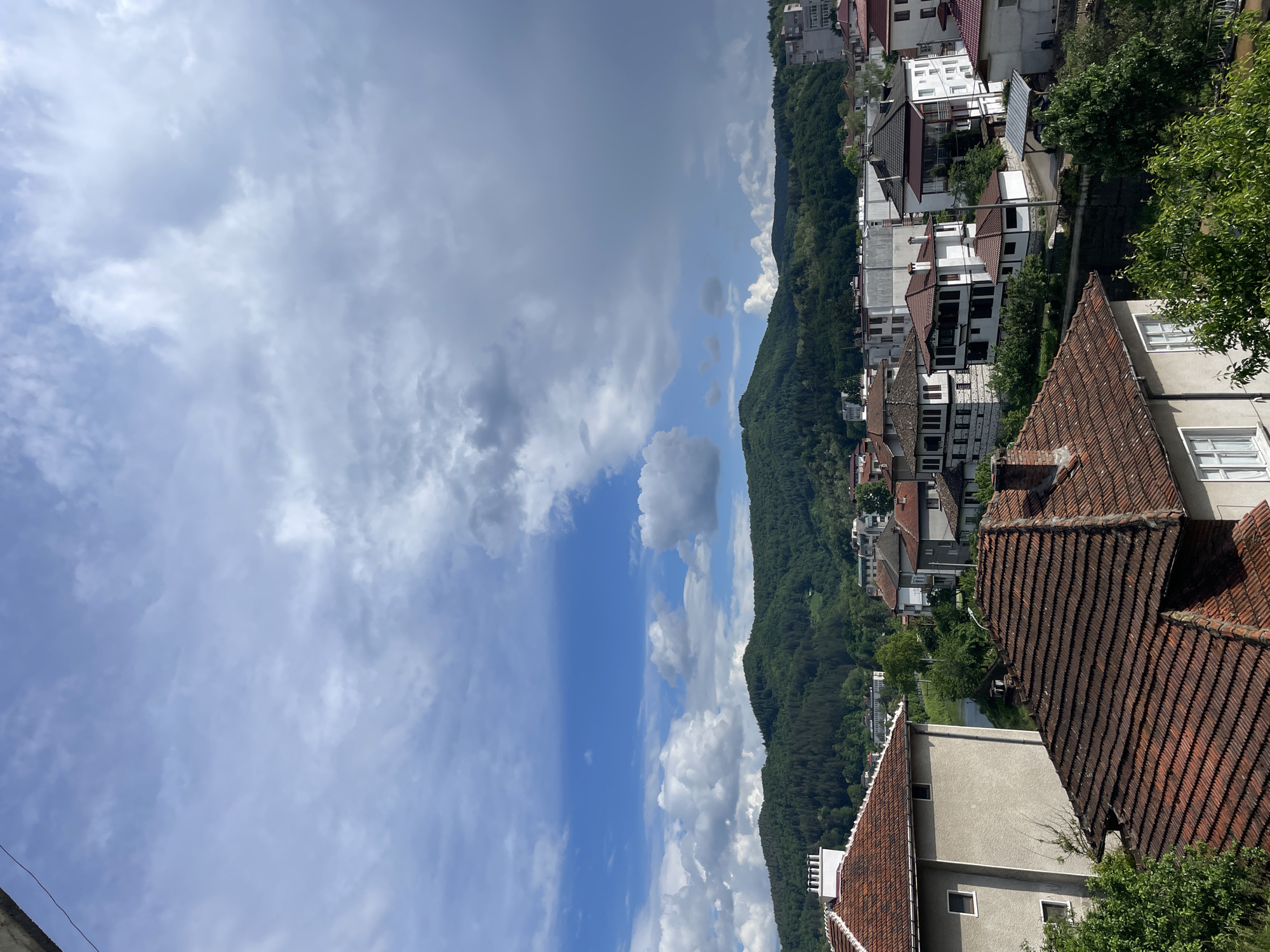 View from an elevated position of Zlatograd, Bulgaria. The scene features a cityscape nestled along a tree-covered mountain slope. Buildings with red-tiled roofs and white facades are prominent. The sky is filled with mixed clouds, showing both overcast and sunny patches.