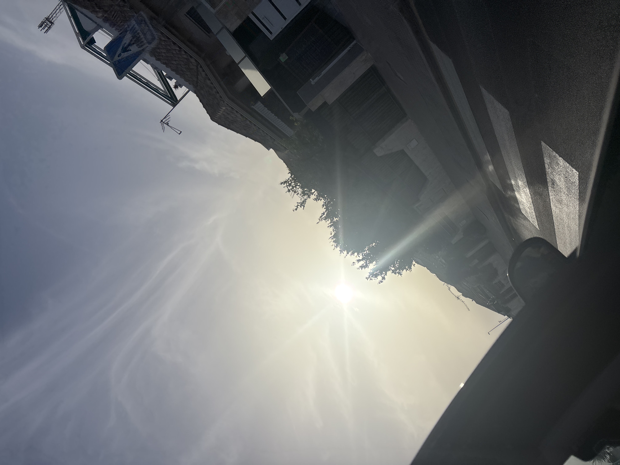 The image depicts a street scene in Orihuela, Spain, angled upwards towards a bright, sunlit sky with thin, wispy clouds. A bright sunbeam emanates from the left side of the frame, causing lens flare and washing out parts of the sky. In the background, residential buildings with terracotta roofs line the street. One building features a prominent blue pedestrian crossing sign mounted on a green metal structure. A street lamp is visible in the distance. In the foreground, a dark object, likely the interior of a vehicle, occupies the lower left portion of the frame, obscuring the view. A wet crosswalk is visible on the asphalt road, reflecting the sunlight. Trees with sparse foliage are also present, casting shadows on the buildings and road.