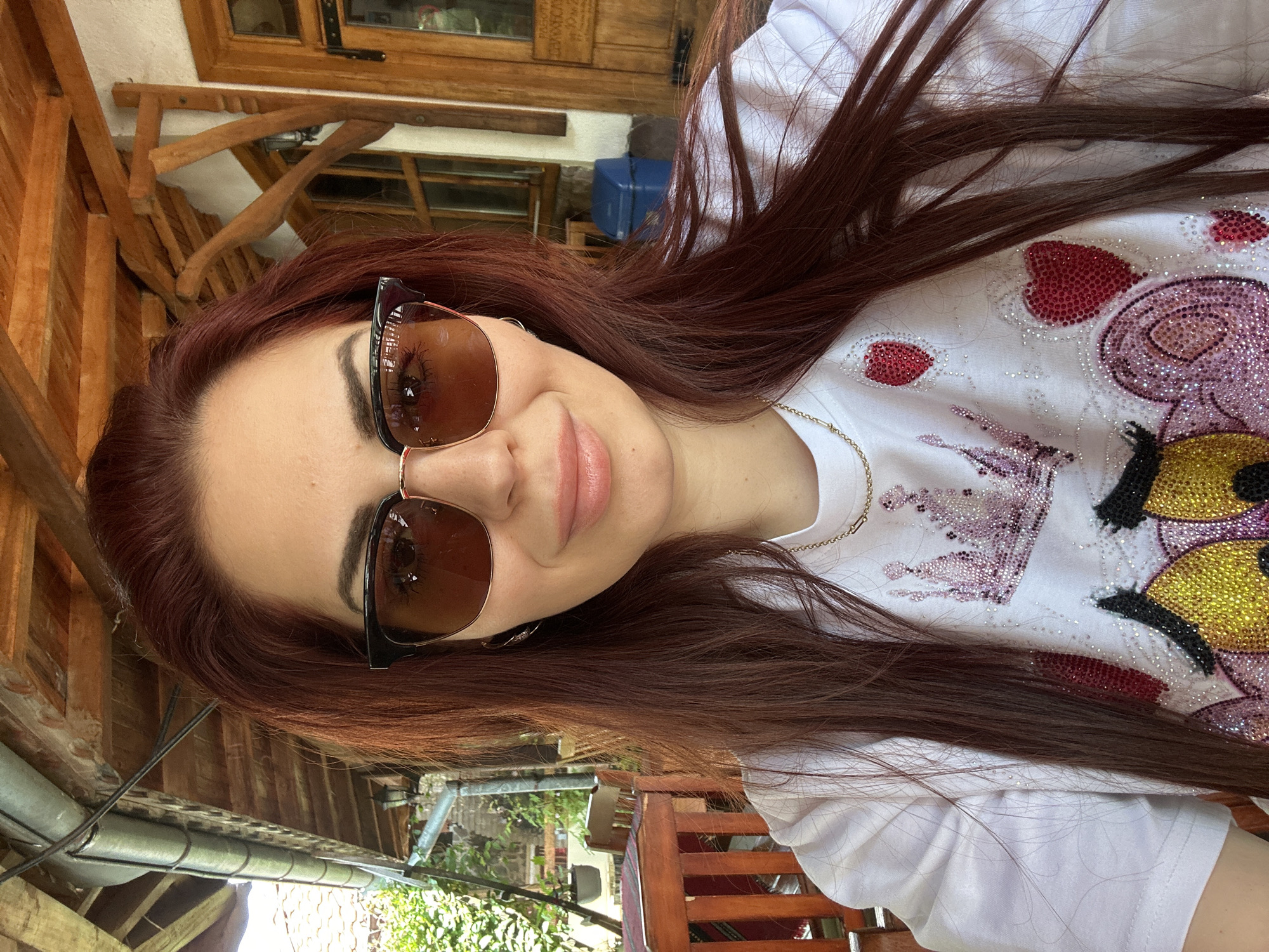 A woman with reddish-brown hair, wearing sunglasses and a white t-shirt with an image of a cartoon character with hearts and gold embellishments, is photographed outdoors, likely in Barziya, Bulgaria. She has fair skin and appears to be wearing lip gloss. A thin gold chain necklace is visible around her neck. The background includes wooden structures suggesting a patio or outdoor seating area, along with glimpses of greenery and a building in the distance. A drainpipe is visible in the foreground, indicating an exterior location.