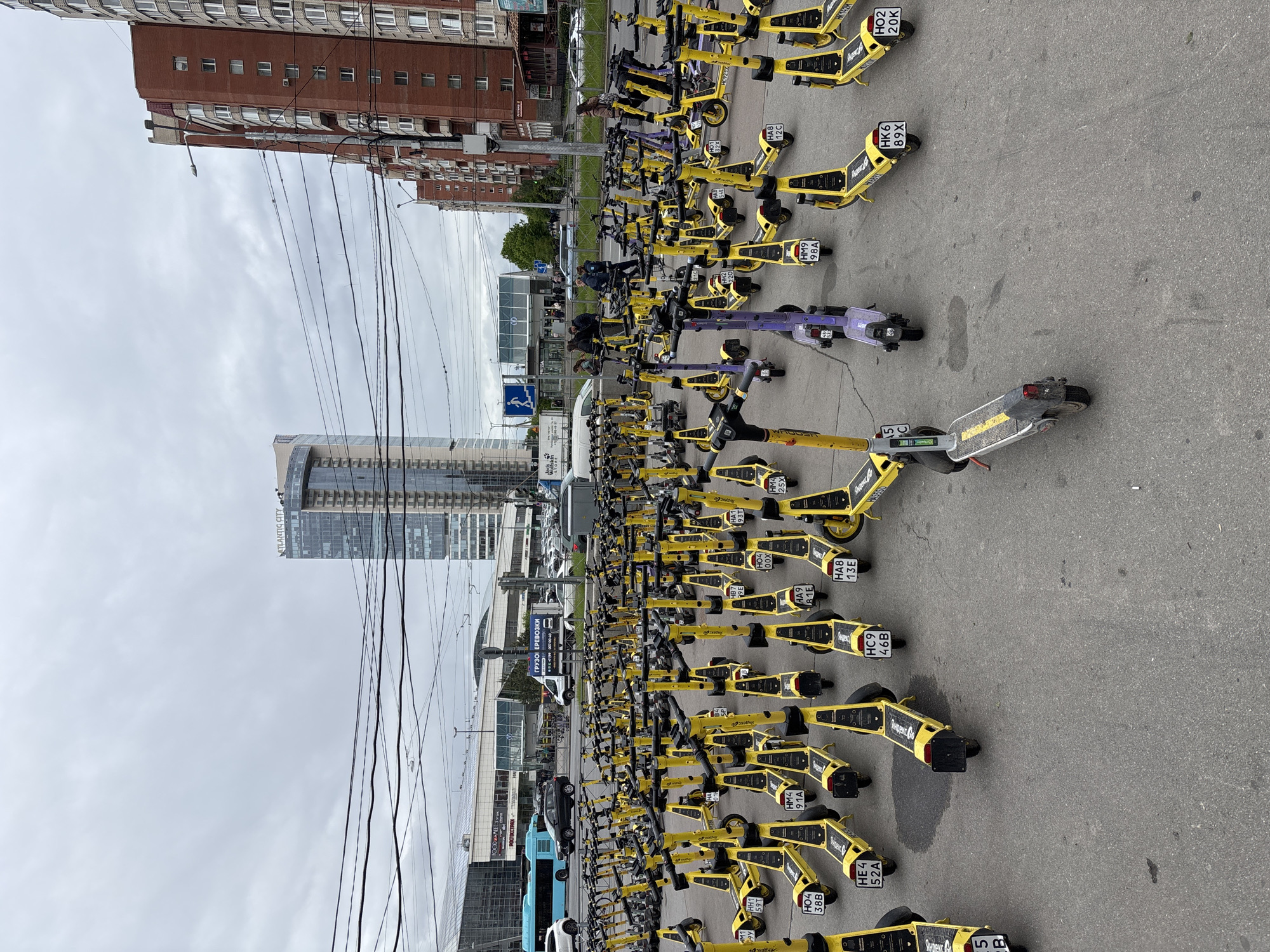 The image shows a large number of rental electric scooters parked on an asphalt surface in Saint Petersburg, Russia. The scooters are mostly yellow with some purple and gray ones interspersed. Some scooters have license plates with Cyrillic characters and numbers. In the background are various buildings including a high-rise building labeled "ATLANTIS CITY" and a brick apartment building. Power lines stretch across the sky. A bus and other vehicles are visible in the distance. Some people are present near a building with glass walls. The sky is overcast.