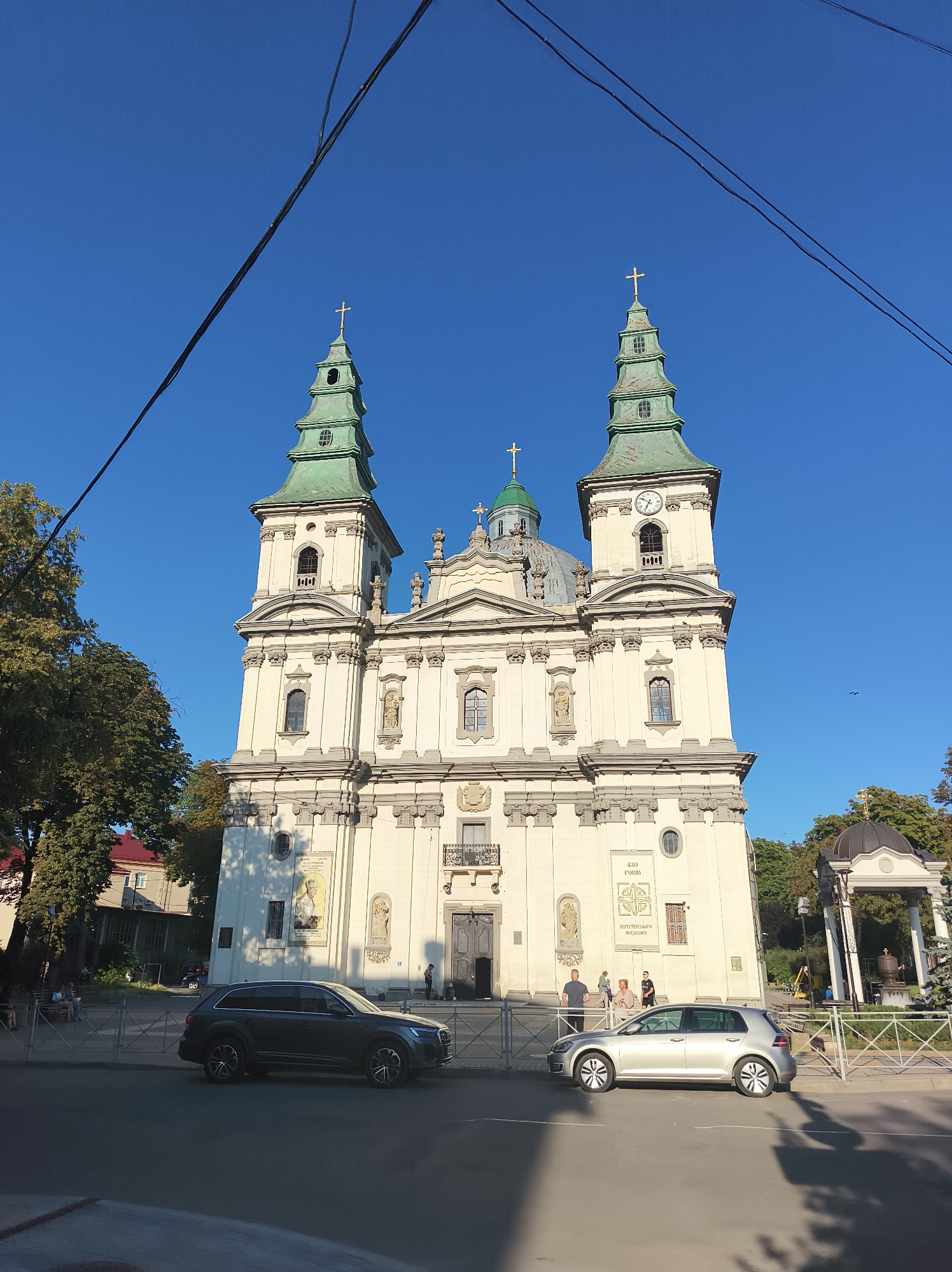 The image shows a large, ornate church with two prominent green-domed towers and a central dome, against a clear blue sky. The church is a light color, likely stone or stucco, and features detailed architectural elements like columns, statues, and arched windows. A clock face is visible on one of the towers. In the foreground, two cars are parked on a street in front of a metal fence. One car is a dark SUV, and the other is a silver hatchback. Several people are visible walking and standing near the church and along the sidewalk. Trees with green foliage are present on the left and right sides of the church, suggesting an urban setting with some greenery. Power lines are strung across the top of the image. This is the Ternopil Cathedral, also known as the Cathedral of the Immaculate Conception of the Blessed Virgin Mary, located in Ternopil, Ukraine.