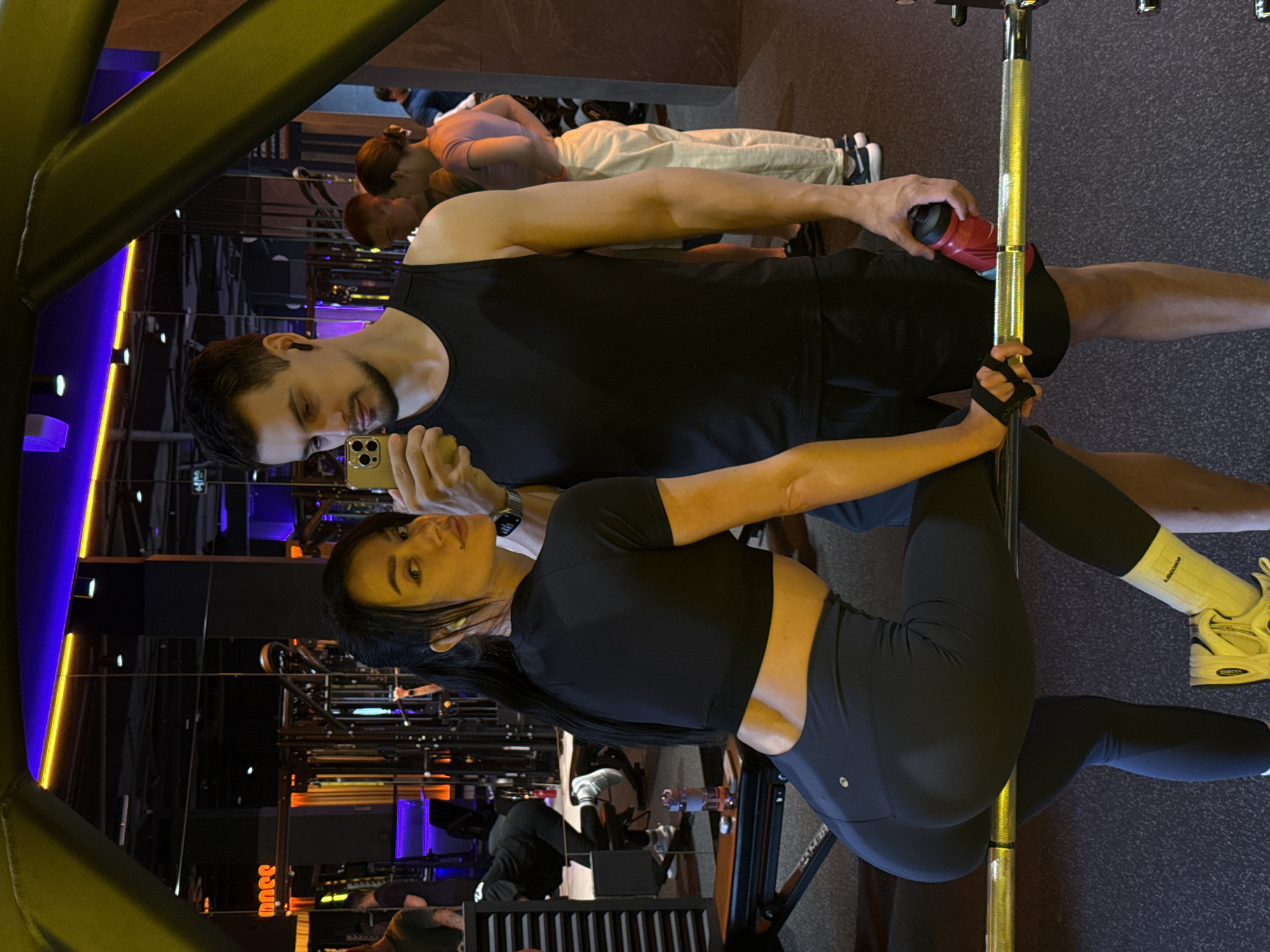 Here's a description of the image:
The image depicts a couple taking a selfie in a gym. The man is holding a phone, and both are looking towards the camera. The woman is leaning against a vertical bar. She is wearing a black crop top and leggings, and the man is wearing a black tank top and shorts. The gym has various exercise equipment visible in the background. In the background there are also other gym-goers using the equipment. A water bottle sits on a ledge near the man. The location is Krasnoyarsk, Russia.
