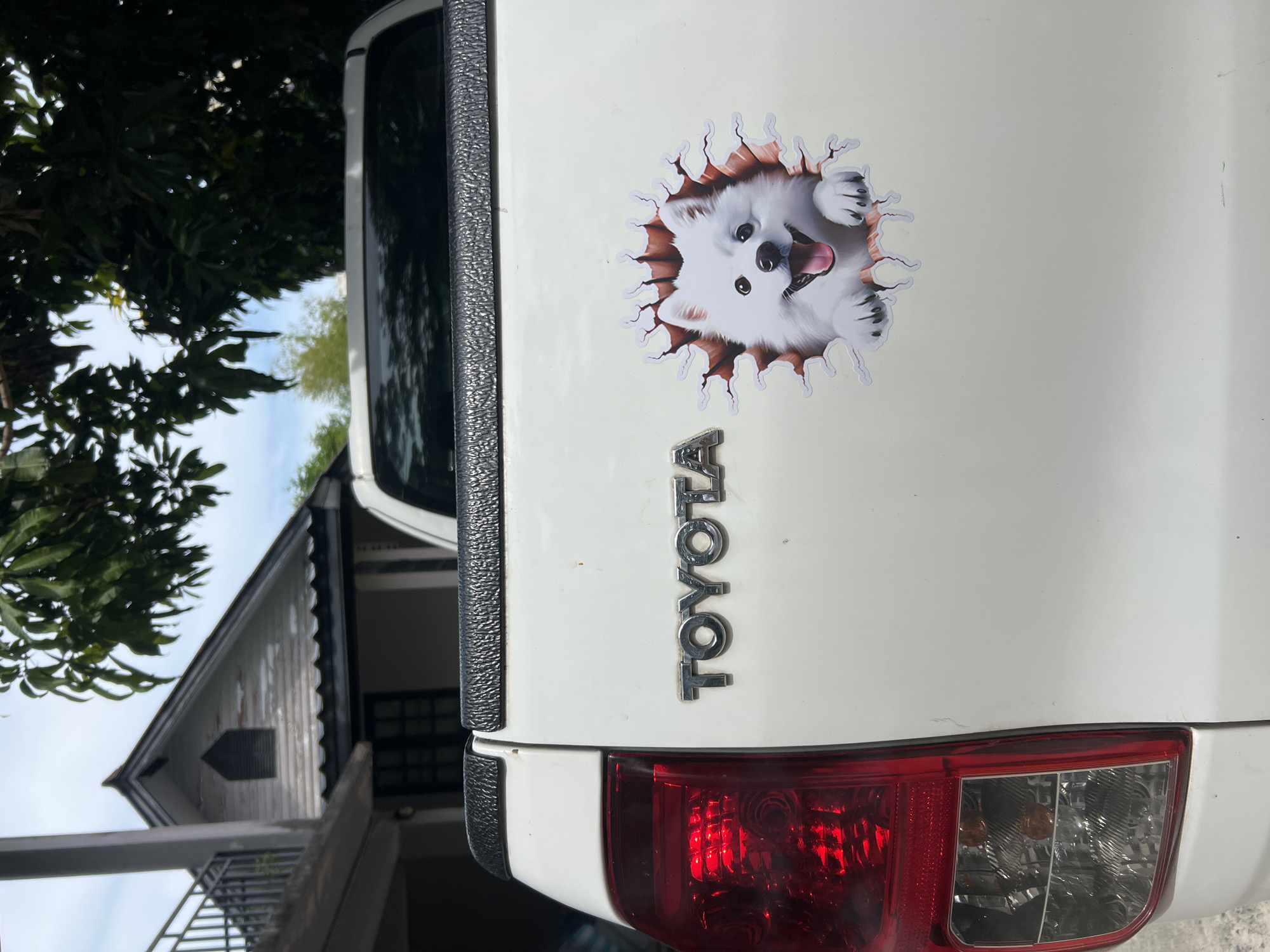The image shows the rear of a white Toyota pickup truck. The Toyota logo is affixed vertically on the tailgate. A dog sticker is also present on the tailgate, above the logo. In the background, there's a house with a slanted roof and green foliage. The location is Baan Lamai, Thailand.