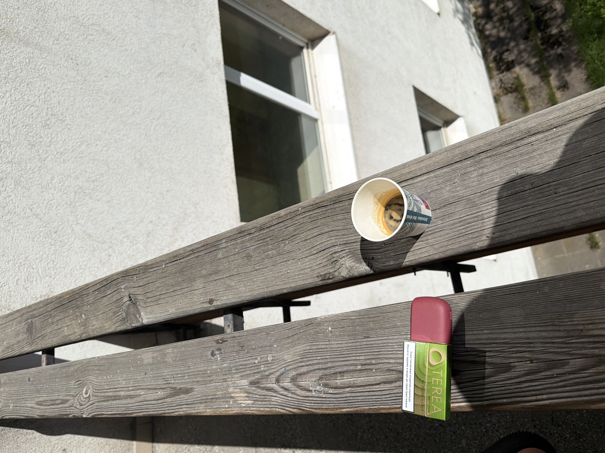 The image depicts an outdoor scene, viewed from a slightly elevated, oblique angle, focusing on a weathered wooden bench. On the bench, an empty, disposable coffee cup and a TEREA tobacco heating product package are prominently displayed. The white coffee cup, adorned with blue and red patterns, holds dark coffee residue and features the English text "Attention: Hot drink." Next to it, a maroon IQOS ILUMA device rests on its green TEREA SOFT FUSE package. The packaging explicitly states "OTEREA," "SOFT FUSE," and "for IQOS ILUMA." A health warning in Bulgarian is visible on its side: "Това тютюнево изделие уврежда Вашето здраве и води до пристрастяване" (This tobacco product harms your health and is addictive).
The wooden planks of the bench exhibit pronounced grain, knots, and signs of weathering, suggesting age and exposure to the elements. In the background, a light-colored building with a textured, possibly stucco, facade is visible. Two windows with white frames are present, with the larger one on the left reflecting bright sunlight. Beyond the bench and the building, a glimpse of green grass and dirt, dappled with shadows, indicates a sunny day. The strong, defined shadows cast by the objects and the bench suggest it is midday or early afternoon. The scene is devoid of people, conveying a quiet, paused moment, possibly in a courtyard or outdoor seating area in Sofia, Bulgaria, as indicated by the Bulgarian text and provided location context.