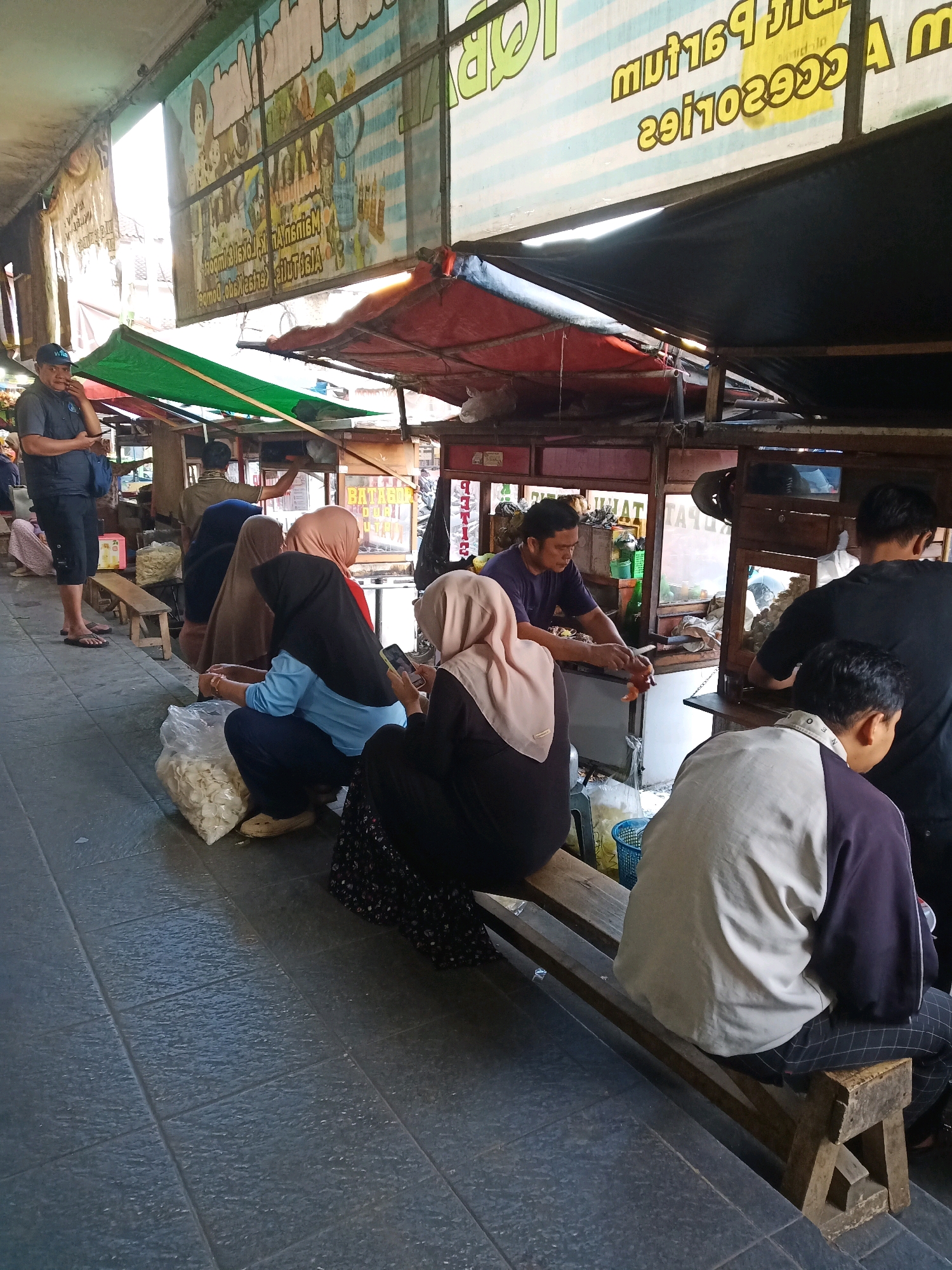 The image depicts an outdoor market scene in Garut, Indonesia. In the foreground, several people are seated on a wooden bench, some facing away from the camera. A woman in a black headscarf and blue shirt is crouching next to a large plastic bag of white snacks. To her right, another woman wearing a light brown headscarf is looking at her phone. Behind them, a man in a dark shirt is cooking at a food stall.
The market stalls are covered by tarps, with one stall displaying a red tarp and another a green tarp. Banners with text in Indonesian are visible hanging above the stalls. The ground is paved with dark gray tiles. The overall atmosphere suggests a busy marketplace where food is being prepared and sold.