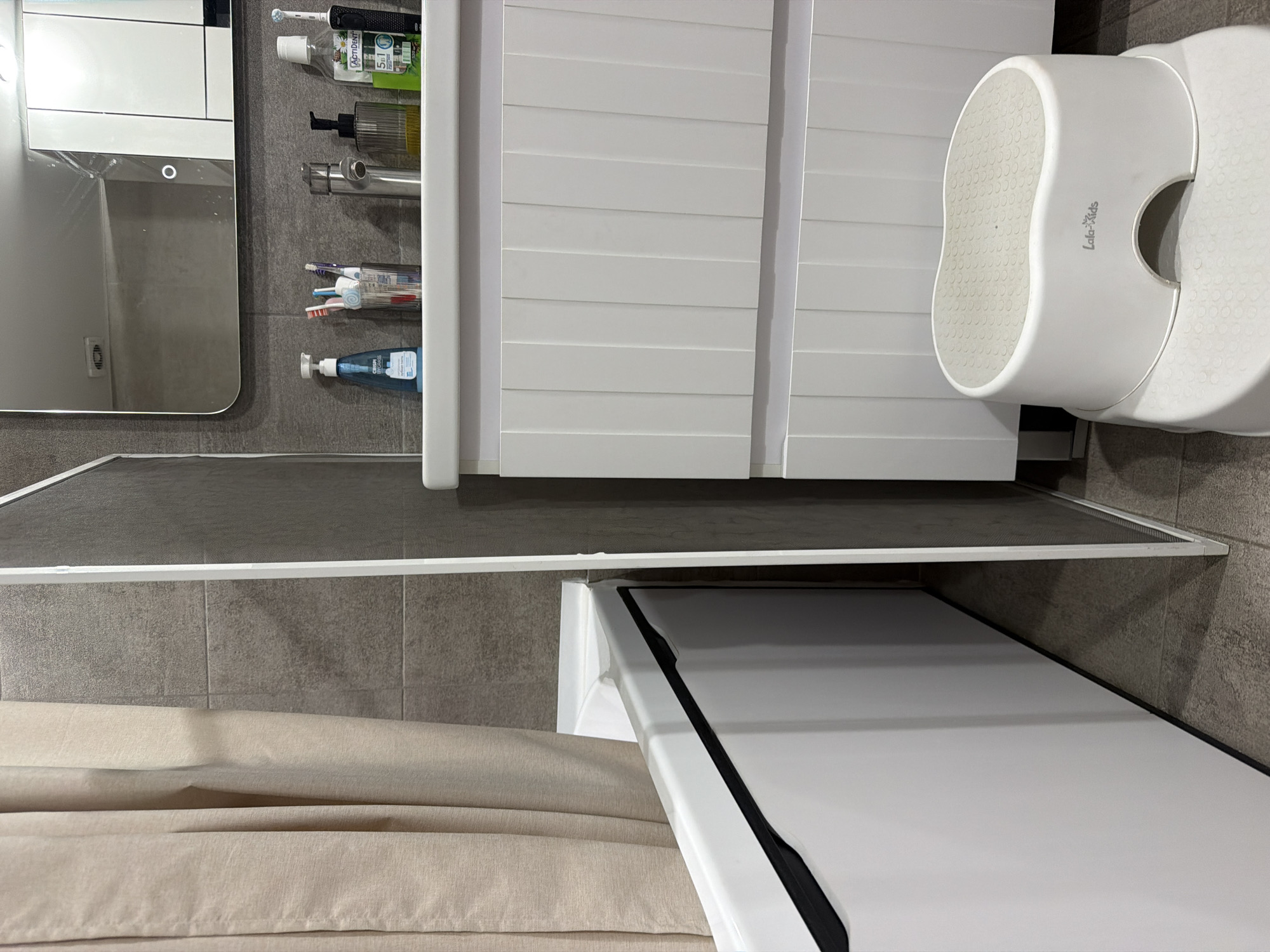 The image depicts a modern bathroom interior in the city of Chaikovsky, Russia. On the left, a beige fabric shower curtain partially drapes over a white bathtub with a prominent black trim. The wall behind the bathtub is tiled with large, rectangular, dark grey tiles. A thin, white vertical partition separates the bathtub area from the vanity on the right.
The right side of the image features a white vanity cabinet with two ribbed drawers, resting on chrome feet. A white countertop supports a collection of toiletries, including a blue pump bottle labeled "CRISPY BY GRASS", a glass holding multiple toothbrushes, a silver faucet, a clear ribbed pump bottle containing dark liquid, a bottle of "ACTIDENT 5in1" mouthwash, and a black electric toothbrush. Above the vanity is a wall-mounted, rectangular mirror with rounded corners, featuring a circular illumination point at its base. The mirror reflects a ceiling vent and parts of a white cabinet structure.
Below the vanity, two white plastic step stools are stacked, with the smaller top stool bearing "Lala-Kids" branding and a light grey textured surface. The floor is tiled with large, dark grey tiles, matching the wall tiles. No people are visible in the scene.