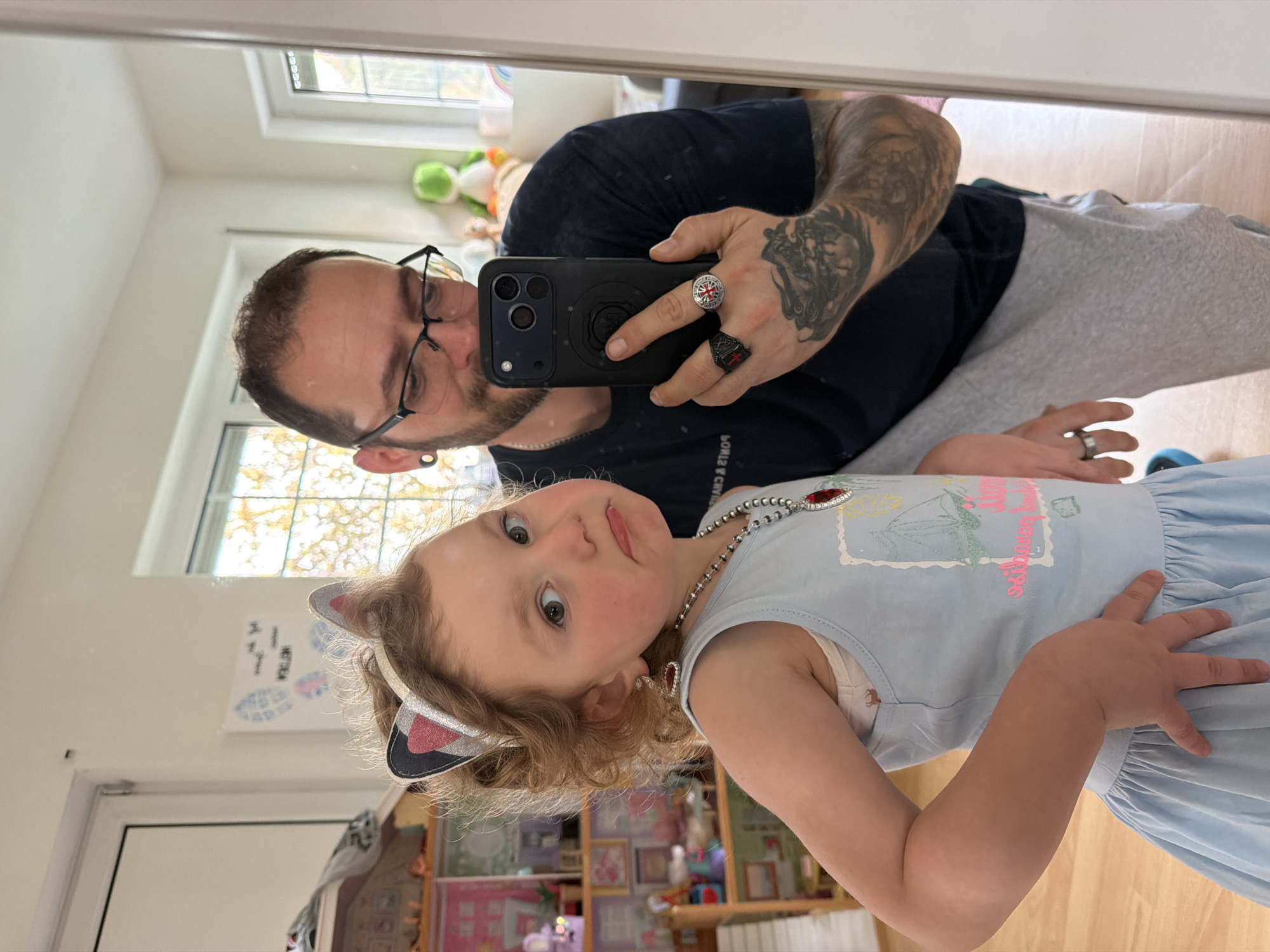 A selfie-style photograph shows an adult and a child in an indoor setting, likely a home. The adult, possibly male, is holding a phone with a triple-lens camera in his right hand, presumably to take the photo in a mirror. He is wearing glasses and a dark-colored T-shirt, along with light-colored sweatpants or similar casual wear. His arms are heavily tattooed, and he is wearing multiple rings on his right hand.
The child, possibly female, is positioned in front of the adult and is wearing a light blue dress and a headband with cat ears. She also has a pearl necklace with what appears to be decorative red pendants.
In the background, there is a window letting in natural light, and various items on shelves, suggesting a child's room or a playroom. Other details, such as artwork on the wall and additional objects on the shelves, are visible but not sharply defined.
There is no clear indication of the image being set in Trassem, Germany.