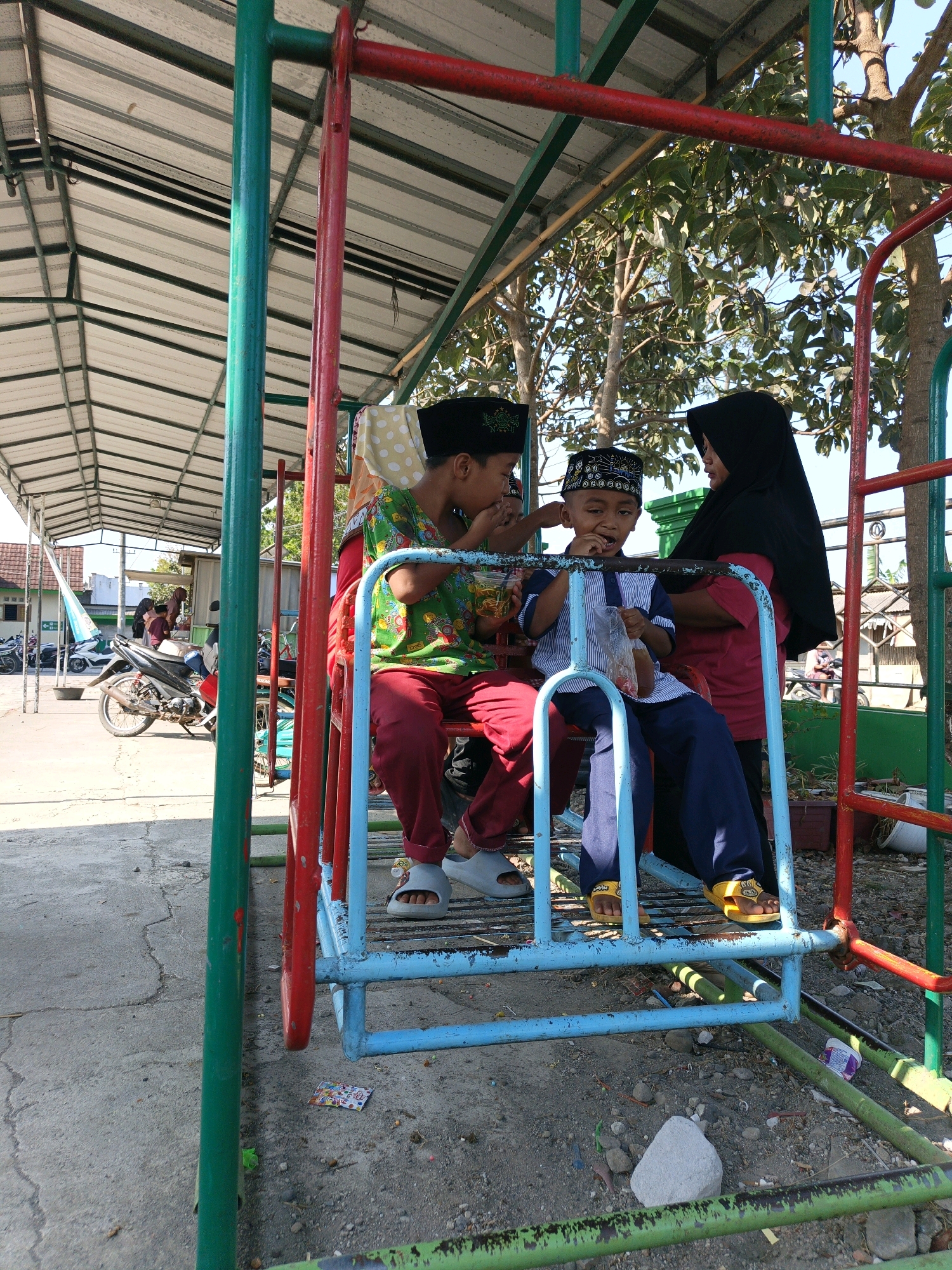 In the city of Kalipurwo, Indonesia, a group of three children and one adult are seated on a blue and red swing set. The children are wearing traditional Islamic attire, with two boys wearing peci hats and one girl wearing a hijab. One of the boys is eating from a plastic bag, while the other two children are looking at something to their left. The adult, a woman wearing a black hijab, is standing behind the children. The swing set is located outdoors, under a covered walkway, with trees and other buildings visible in the background. The ground is paved with concrete and scattered with small rocks and debris. A motorcycle is parked in the background, suggesting a public or school setting.