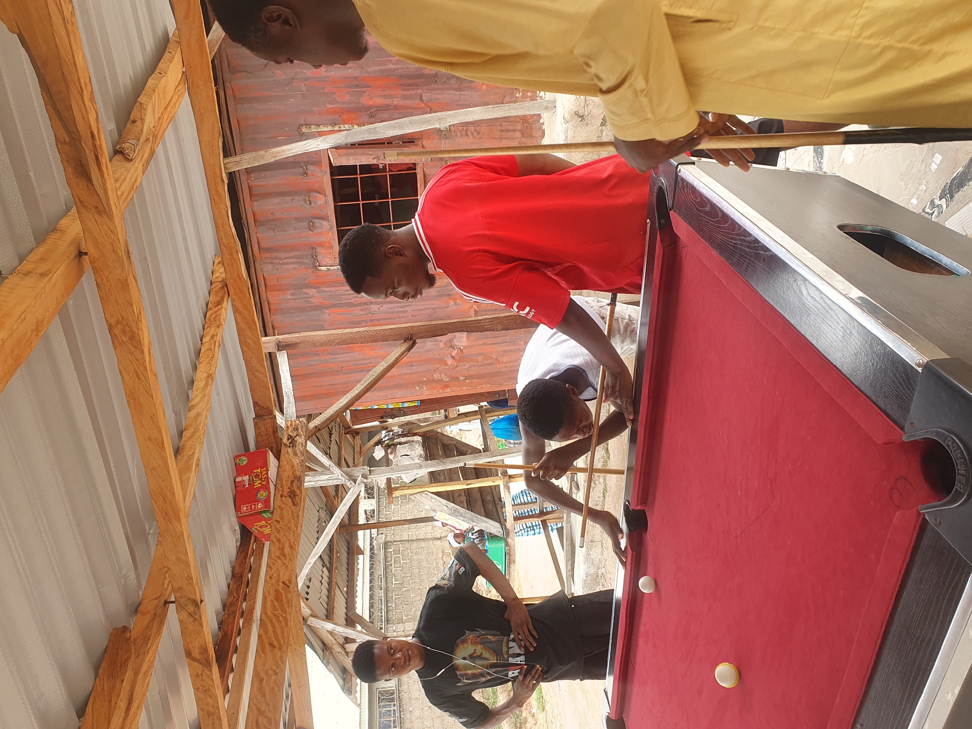 In Lokoja, Nigeria, an outdoor billiards game is underway beneath a rustic, open-air shelter constructed with wooden beams and corrugated metal roofing. Three young African men are actively engaged with a red felt pool table. One man, wearing a black t-shirt with a graphic print and earbuds, stands with his hands on his hips, observing the game. Another man, dressed in a red t-shirt, is positioned across the table. The third participant, in a white t-shirt, is in the process of taking a shot, leaning over the table with a pool cue in hand. Two white pool balls are visible on the table. In the background, a rusty corrugated metal container structure serves as a backdrop, with a wooden staircase and various other structures suggesting a bustling, informal environment.