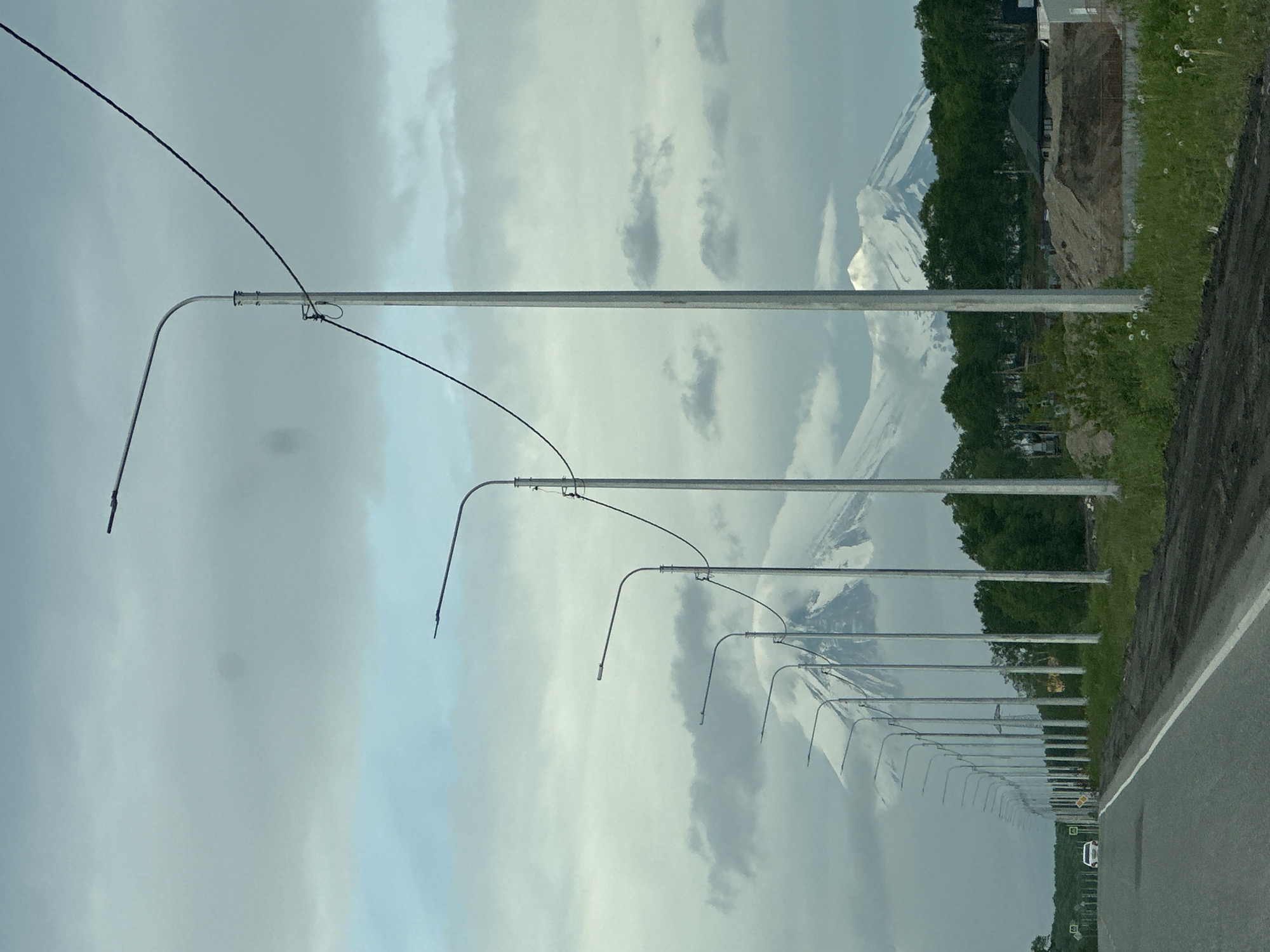 The image depicts a road lined with streetlights leading towards a snow-capped mountain range partially obscured by clouds. The road is paved with a white line marking the lane. On the right side of the road, there is a construction site with piles of dirt and some newly built structures. A line of trees separates the construction site from the mountains. In the distance, on the road, a white car is visible. The sky is cloudy with some patches of blue. The scene appears to be in an outdoor, possibly rural or suburban, setting. The location is identified as Kruetoberegovy, Russia.