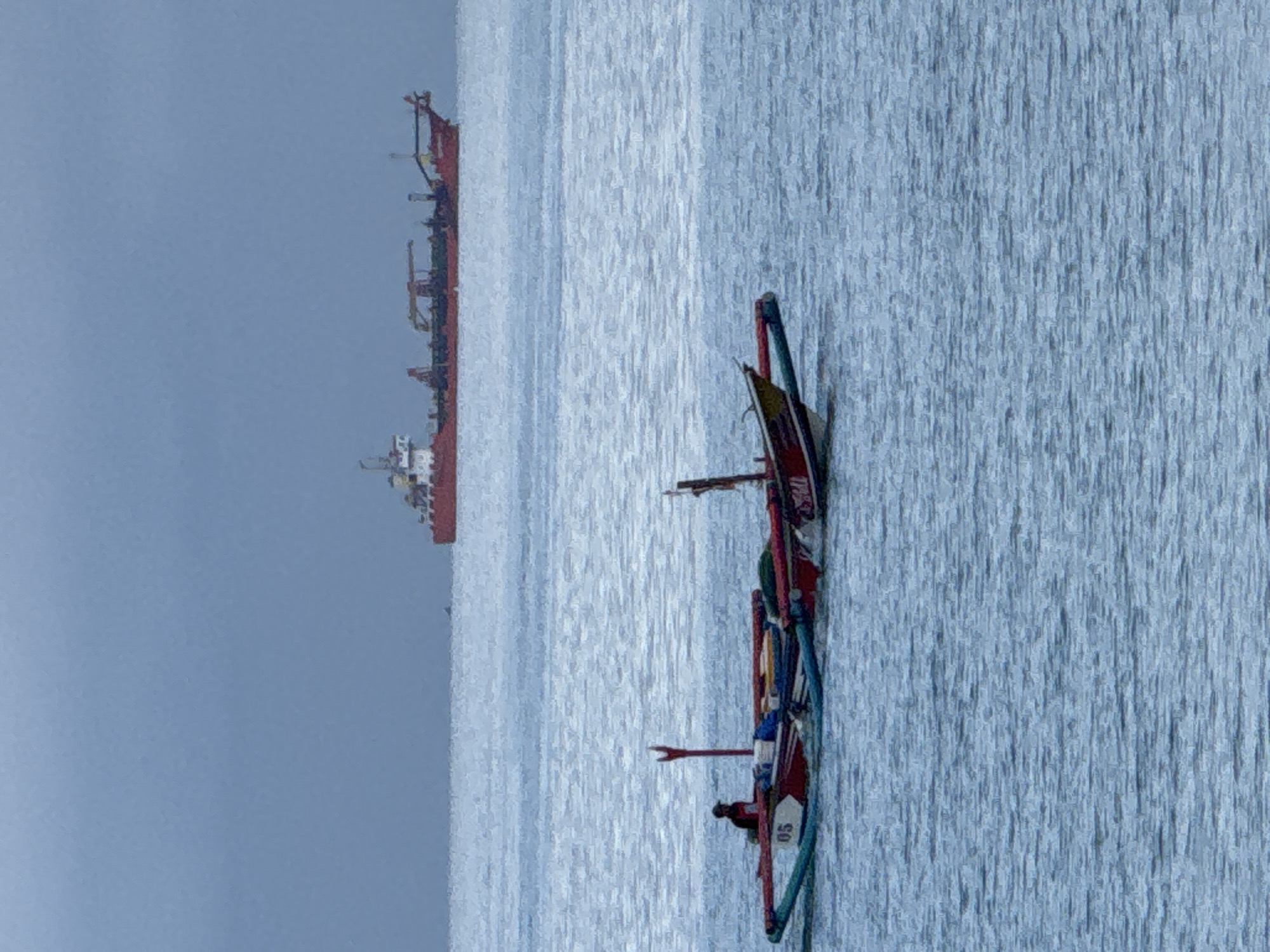 The original image, when viewed correctly oriented, depicts a serene maritime scene under an overcast sky. In the foreground, positioned on the right side, a traditional outrigger boat, likely an Indonesian Jukung, floats on calm waters. This boat is distinctively colorful, painted predominantly in red and white, with striking green and blue accents on its outriggers. A lone figure, visible as a dark silhouette, is seated within the boat, suggesting an individual engaged in fishing or local transit. Visible text on the boat's side includes the number "05" in white, and partially obscured red lettering that appears to read "URES".
In the mid-ground, further to the left, a large red-hulled cargo or tanker ship is visible. This substantial vessel features multiple decks and structures, indicating its industrial scale. It appears to be either anchored or moving slowly across the water. The expanse of water separating the two vessels is largely calm, displaying subtle ripples that reflect the soft, diffused light. The sky above is a uniform light blue-gray, suggesting an overcast day or the subdued light of dawn or dusk. The overall atmosphere is tranquil, devoid of explicit emotions. This scene captures a blend of traditional local seafaring and modern shipping activities in the waters near Kuta Selatan, Indonesia.