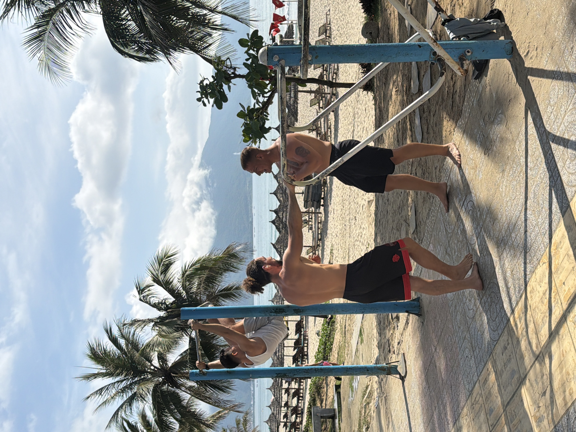 Three shirtless men engage in physical exercise on parallel bars. One man in black shorts performs push-ups; another in black shorts with red trim stands and stretches. The third man, wearing a white tank top, is upside down, gripping the bars. Palm trees frame the image against a backdrop of beach umbrellas, a sandy beach, and the ocean. The blue sky contains scattered clouds. The location is likely a beach park in Đà Nẵng, Vietnam. The surface beneath the bars is paved with patterned tiles.