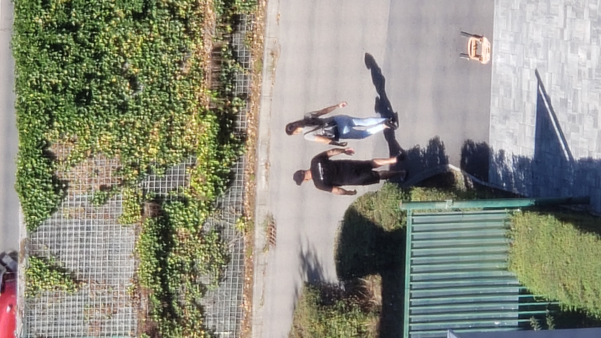 A medium-shot, aerial view captures two individuals walking on a paved surface. The person on the left, a male, wears a black t-shirt, black shorts, and a black cap. The person on the right, a female, is dressed in a grey t-shirt and light blue pants, carrying a dark bag over their shoulder. They are walking away from the camera, casting long shadows on the ground. The environment includes a wire-mesh wall covered in dense green ivy, a paved road with debris, a green metal fence bordering a manicured hedge, and a textured tiled area. The scene appears to be in an outdoor, urban setting. The presence of a small, orange, overturned chair on the tiled surface suggests a recent disturbance or an abandoned item. This location is a section of sidewalk or walkway adjacent to residential buildings or public spaces within the city of Prešov, Slovakia.