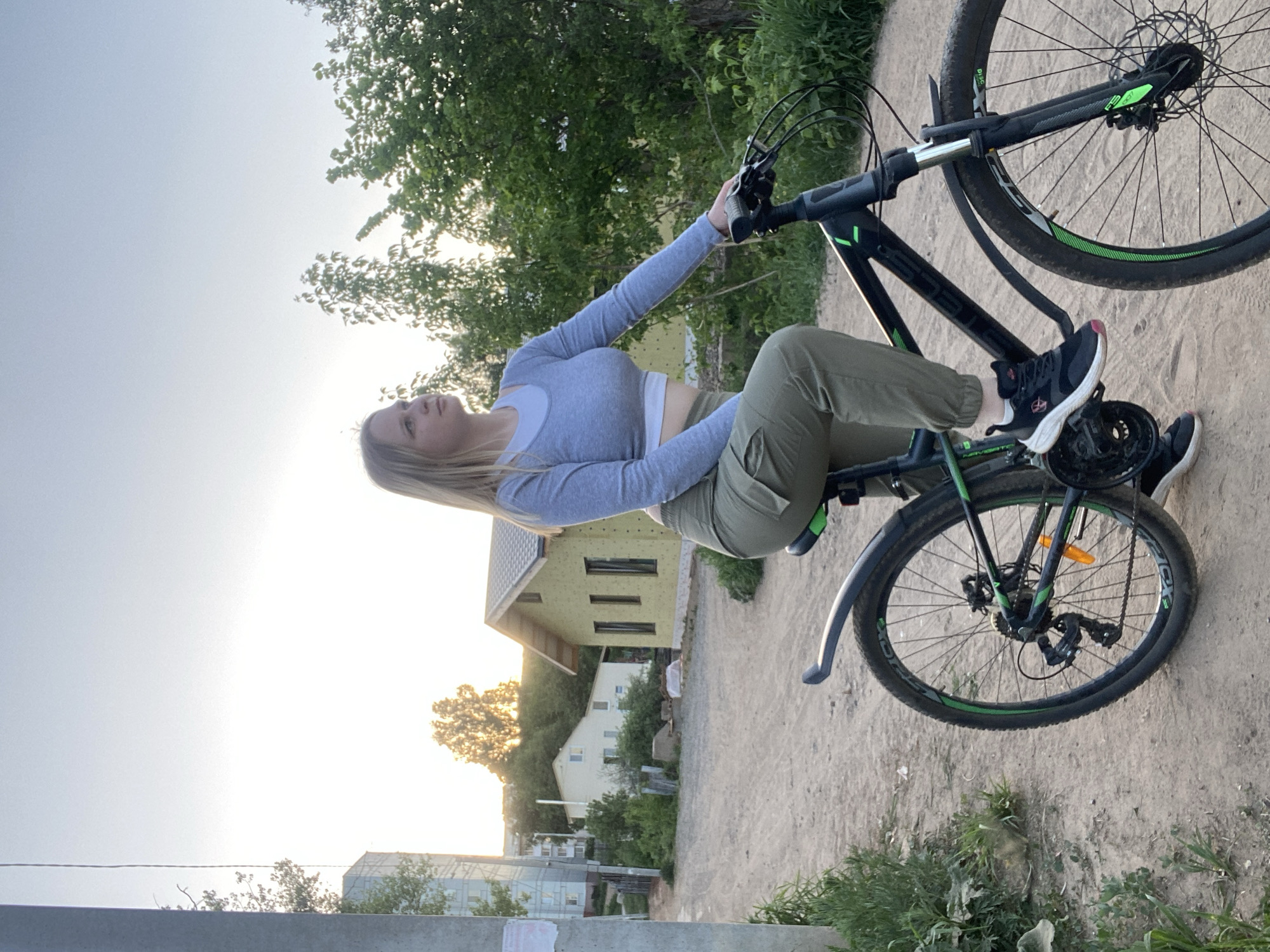 A fair-skinned woman with long blonde hair is sitting on a black and green bicycle in what appears to be a residential area in Chaikovsky, Russia. She is wearing a long-sleeved grey top and olive green pants, with black and pink athletic shoes. She is turned to her left, looking towards the setting sun. Behind her, there is a house under construction with yellow cladding, along with other residences further in the background. The setting is a dirt road with some vegetation bordering it. A concrete pillar is visible in the bottom left corner of the image.