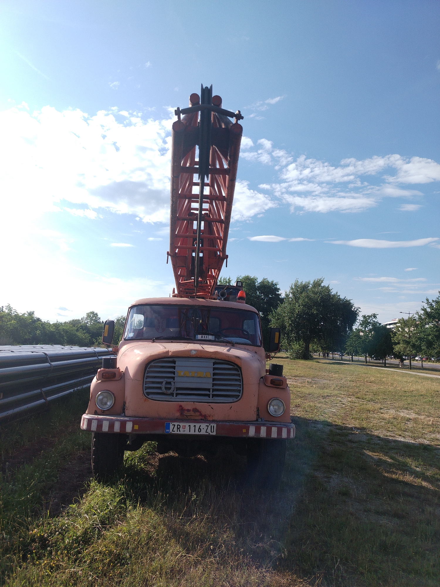 The image shows a front view of an old, orange Tatra fire truck parked on a grassy area. The truck has a large, red, extendable ladder mounted on top, which reaches high into the blue sky with scattered white clouds. The front of the truck features a silver grille with the "TATRA" logo visible, a single round headlight on each side, and a red and white striped bumper. A license plate with the text "ZR 116-ZU" is mounted below the bumper. The scene appears to be outdoors during the daytime, with sunlight casting shadows and illuminating the truck. To the left of the truck, a stack of large, dark, cylindrical objects, possibly pipes, is visible. In the background, there are trees and a road with some buildings and parked cars, suggesting an urban or suburban setting. The overall atmosphere is one of a still, sunny day. No people are visible in the image.