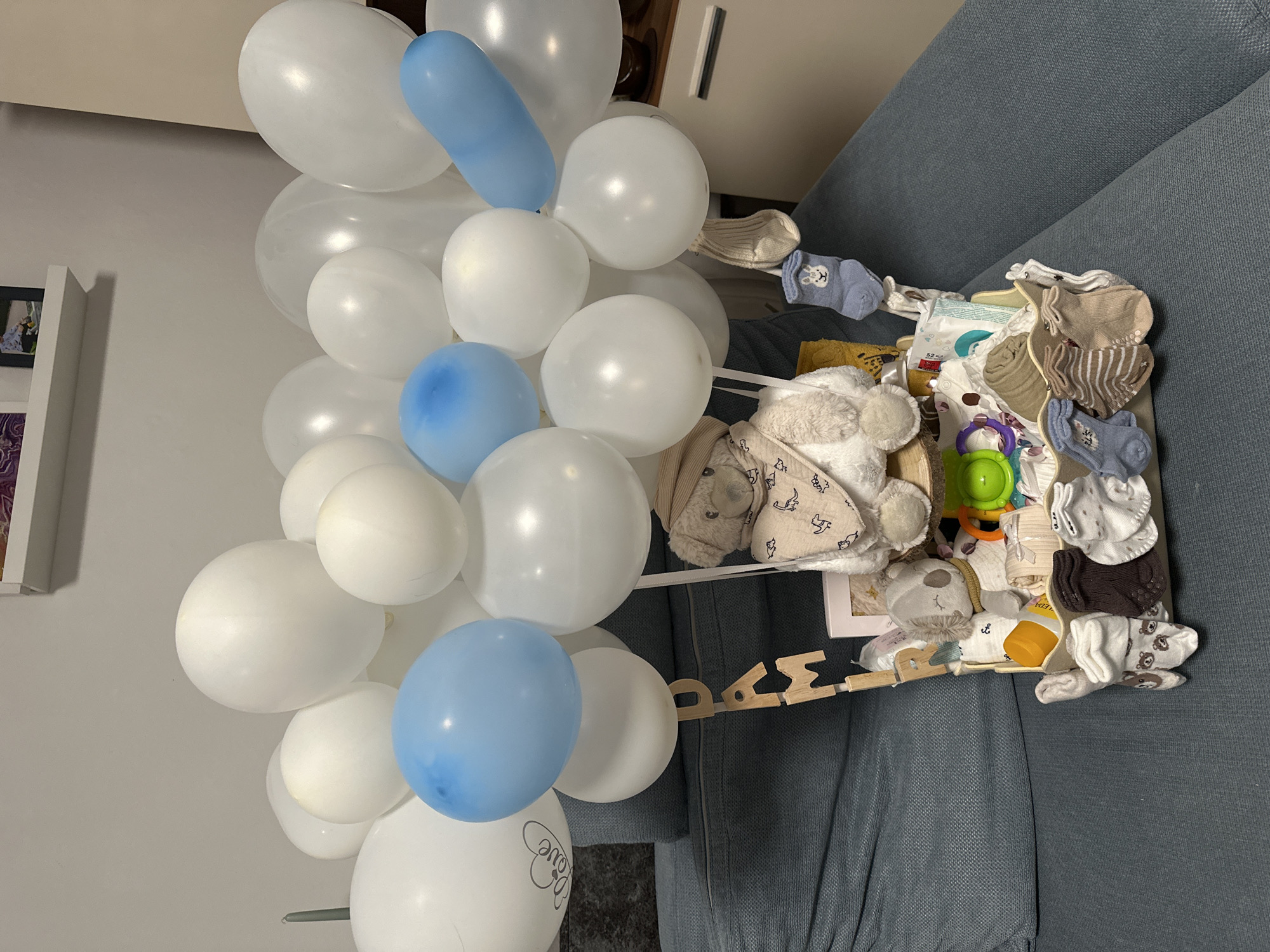 The image shows a baby gift basket designed to look like a hot air balloon, placed on a blue upholstered sofa. The balloon portion consists of numerous white and light blue balloons of various sizes, with a large white balloon on the left featuring a heart and the word "Love" in black lettering. The basket itself, resembling a gondola, is filled with baby items including plush toys (a teddy bear and a koala bear), baby clothes, socks with various patterns, and a small bottle of baby lotion. Wooden letters spelling "DAM" are attached to a wooden support structure for the balloon. In the background, a white shelf with framed pictures is visible on a light-colored wall. The scene appears to be indoors, likely in a residential setting, and there are no people present in the image. The lighting suggests an indoor artificial light source. The specific location in the city of Bad Endorf, Germany cannot be determined from the image itself.
