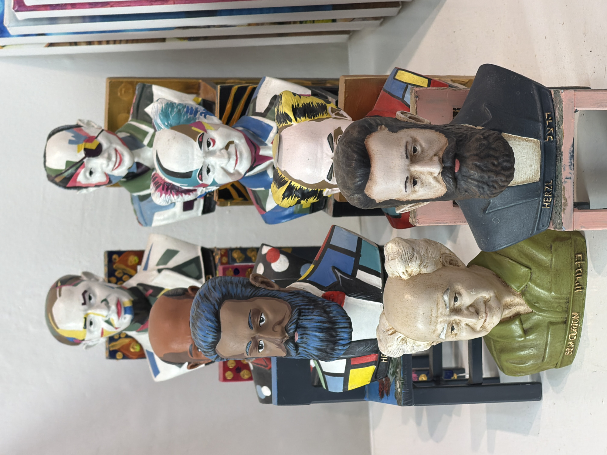 The image captures a display of painted busts, likely made of plaster or similar material, arranged in two rows. The busts vary in depiction, with some appearing abstract and geometric, and others more realistic portraits of historical figures, identifiable by inscriptions on their bases, which include Theodor Herzl and Ben Gurion. The inscriptions are in both English and Hebrew. The style of painting across the busts is colorful and somewhat whimsical.
Given the presence of figures prominent in Zionist history, and the location tag of Tel-Aviv, Israel, it is reasonable to assume this display is located in a museum, gallery, or cultural center relating to Israeli history or art within Tel-Aviv. The environment suggests an indoor setting, possibly a display shelf within the aforementioned location. The lighting is bright and even, illuminating the various colors and details of the busts.