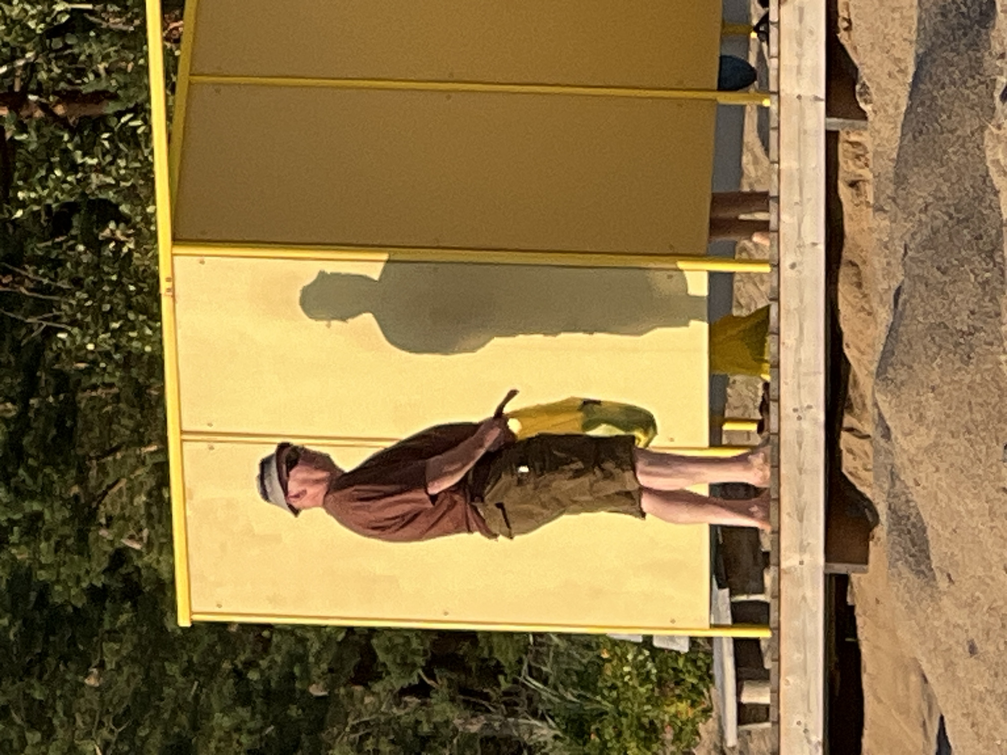 A man stands outdoors in front of a yellow changing room structure, partially silhouetted by the sun. He is wearing a brown t-shirt, olive green shorts, and a bucket hat, with sunglasses on his face. He is holding a yellow and green folded object in his right hand. The man is standing on a wooden platform above a sandy area with some vegetation in the background. Another person's shadow is visible on the yellow wall to the right of the man. The scene appears to be a public beach or recreation area.