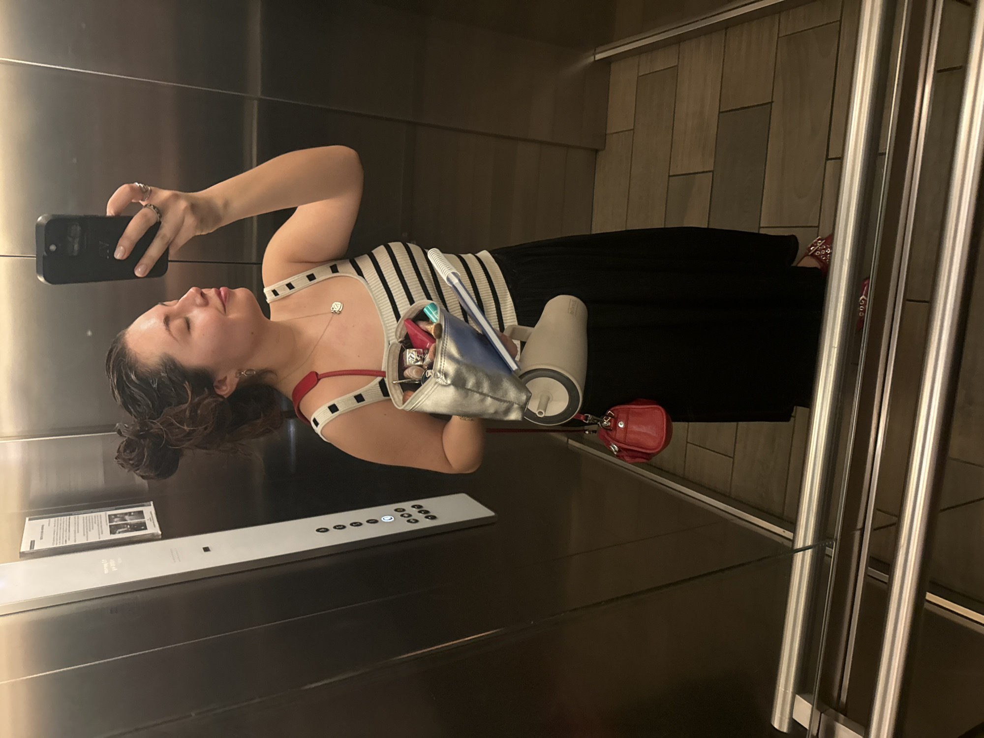 The photograph shows a woman taking a selfie in an elevator. She is holding a smartphone in her right hand, with her arm raised to capture her reflection in the elevator's mirrored wall. She is wearing a black and white striped top, a black skirt, and red shoes. Her hair is styled in a bun.
In her left hand, she holds a silver tote bag containing various items, including what appears to be makeup and a white container. A small red purse is attached to the tote bag.
The elevator's interior features stainless steel walls, a control panel, and a floor covered in light brown tiles. A document is affixed to the wall near the control panel.
The photograph was taken in Mexico City, Mexico.