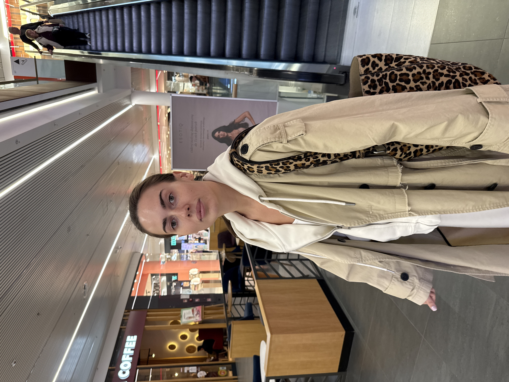 A portrait-oriented photo shows a young woman in the foreground of a multi-level shopping mall in Warsaw, Poland. She looks directly at the camera with a neutral expression. She wears a light beige trench coat over a white hooded top, with a leopard print strap of a bag visible over her shoulder. A matching leopard print handbag is seen resting on the floor or a nearby surface to her right.
In the background, to the left, an escalator ascends with a few people on it. The mall features modern architecture with clean lines and bright, linear lighting fixtures on the ceiling. On the right side of the image, various shops are visible across multiple levels. One shop prominently displays a "COFFEE" sign in white letters on a dark background. Further back, an advertisement banner features a woman and the brand "ESOTIQ" with Polish text that reads: "Odkryj bieliznę dopasowaną do Twoich potrzeb. Otulającą i modelującą sylwetkę" (Discover lingerie tailored to your needs. Hugging and shaping the silhouette). Another shop, likely a cafe, has distinctive circular light fixtures on its interior wall. The setting suggests an active indoor environment during the daytime.
