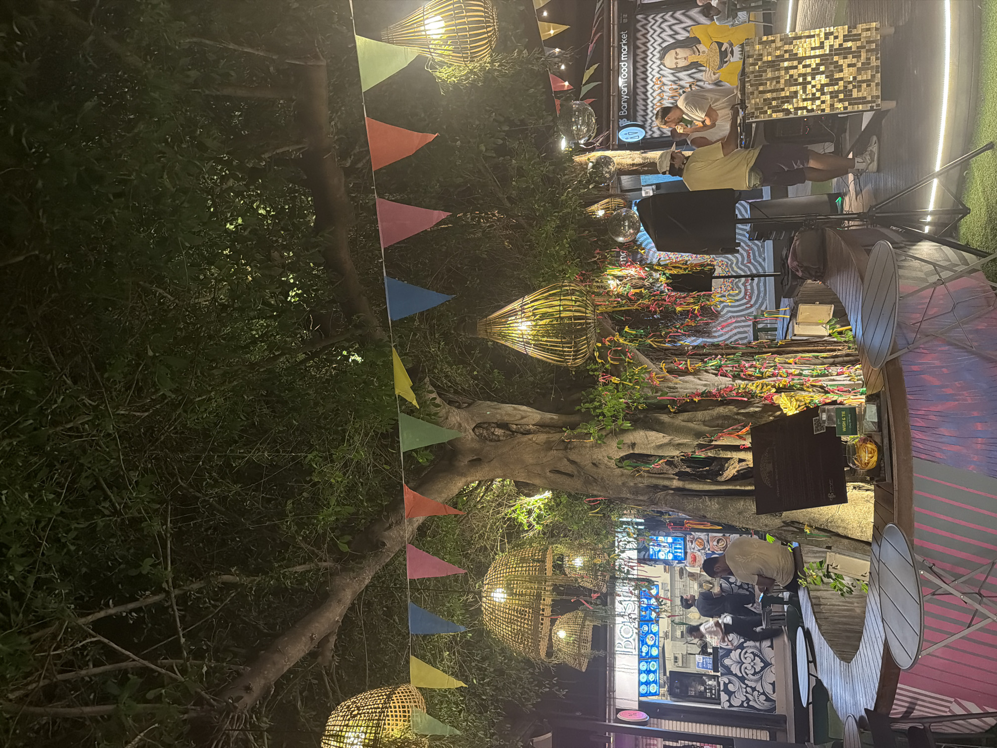 Captured at night in บ้านบางรักษ์, Thailand, the image portrays an outdoor venue, potentially a market or eatery. A massive, illuminated tree dominates the scene, adorned with string lights and decorative flags. Underneath, food stalls are bustling with activity, showing staff attending to customers. Tables and chairs are arranged nearby, catering to diners. Overhead, lamps in woven, basket-like designs cast a warm glow. The environment is active, suggesting an evening crowd.