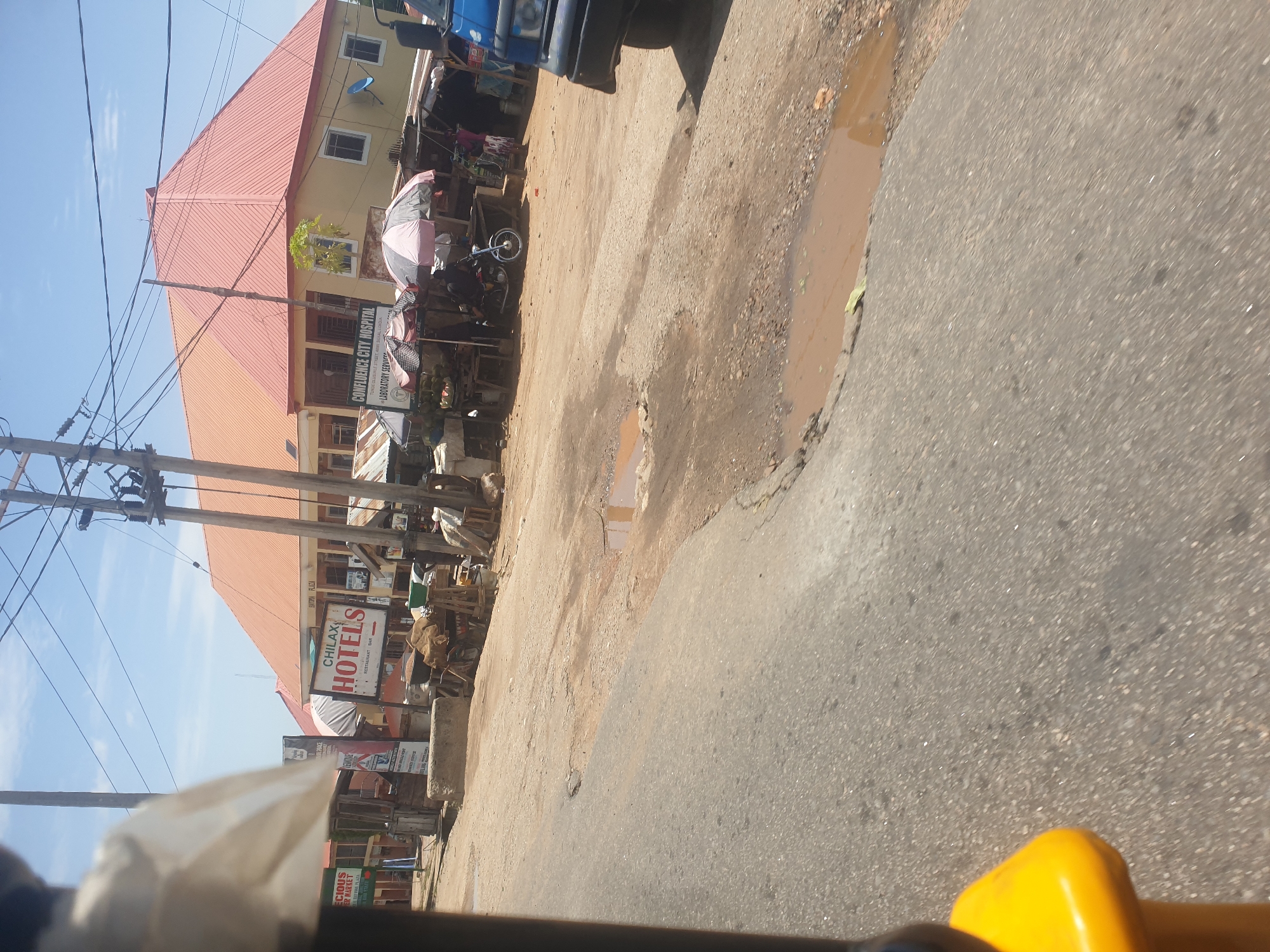 The image depicts a street scene in the city of Unknown, Unknown. The road is partially paved and shows signs of disrepair with potholes and puddles. A building with a red roof is visible on the left, with signs indicating the presence of businesses such as "Chilax Hotels" and "Confidence City Hospital." Several people are present, engaged in activities such as standing near stalls and vending. Utility poles with numerous wires are visible, suggesting a somewhat developed area. A vehicle, possibly a bus or truck, is partially visible in the upper right corner. The image appears to have been taken from a moving vehicle, with the yellow portion of the vehicle visible in the foreground.