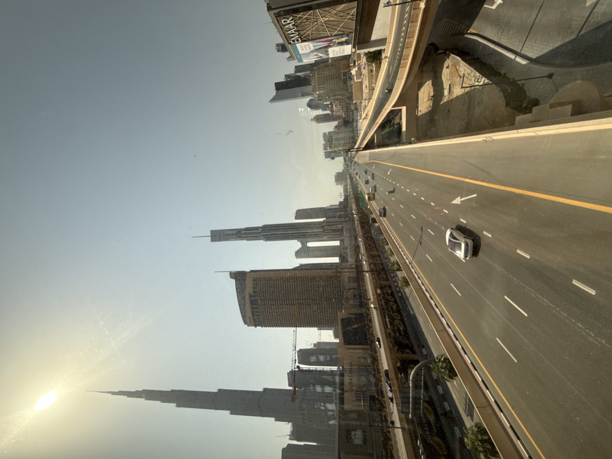 The image captures a wide-angle view of a highway in Dubai, United Arab Emirates, on a clear, sunny day. The Burj Khalifa, the world's tallest building, is prominently visible on the left, silhouetted against the bright sun. The highway, with multiple lanes, is flanked by elevated roadways and futuristic skyscrapers. Several cars are driving on the highway, with one white car in the foreground. Advertisements are visible on the side of the highway, with one large billboard featuring the "EMAAR" logo. The scene depicts a modern, urban landscape characterized by high-rise architecture and well-developed infrastructure.