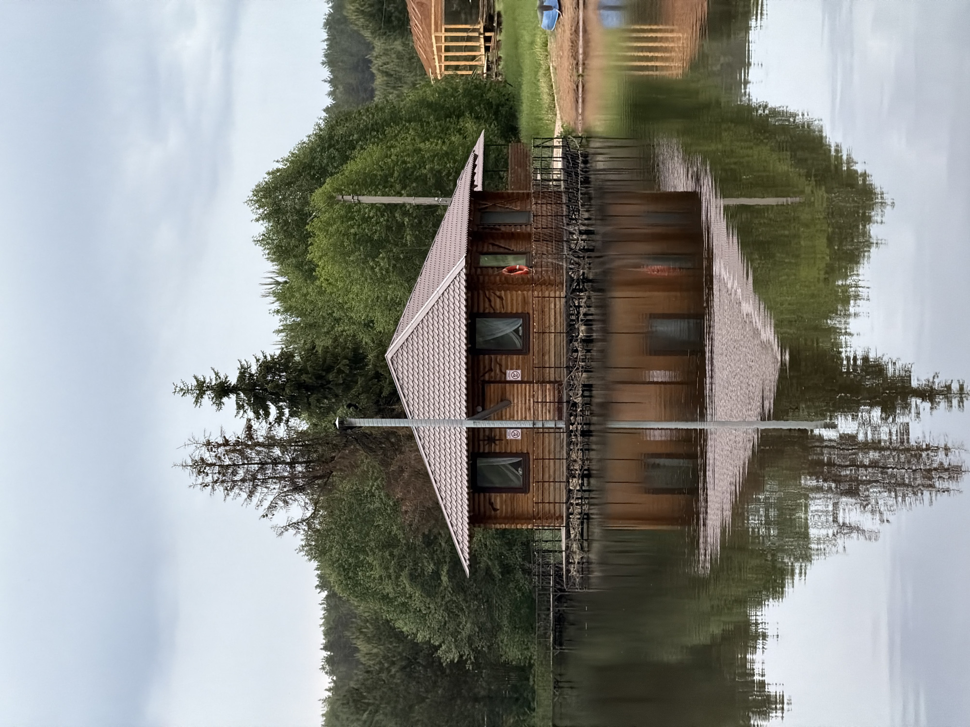 The image depicts a wooden cabin built on stilts over a body of water, likely a lake or river, in Prikamskii, Russia. The cabin has a pitched roof covered with grey tiles. A chimney extends from the roof. Windows are visible on the cabin's facade, and a life preserver hangs on one side. A metal railing surrounds the cabin. The stilts supporting the structure are submerged in the water. The background consists of dense green foliage. The water surface reflects the cabin and surrounding vegetation, creating a symmetrical mirror image. A power pole is visible near the cabin. The sky is overcast. Another wooden structure sits further inland.